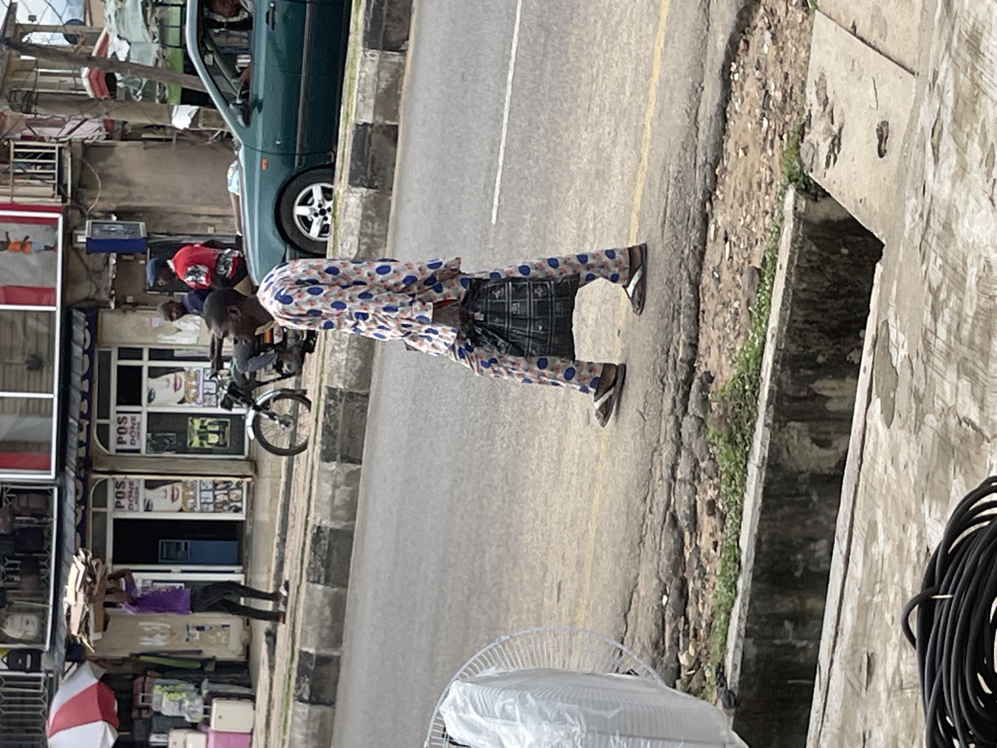 A person in a white, blue, and orange patterned outfit walks down a street. The outfit consists of a long-sleeved shirt and matching pants. The person is wearing flip-flops. The scene is outdoors, with a street in the foreground and shops lining the background. A dark green car is parked on the left side of the street. Other people and objects are visible in the background, including a motorcycle, a red shirt, a purple shirt, and a white and red umbrella. The ground is paved with some dirt and grass visible along the edge of the street. There is a drainage ditch running alongside the road. The lighting suggests it is daytime, with no indication of weather conditions. Some text is visible on the shop windows, including "POS DONE".