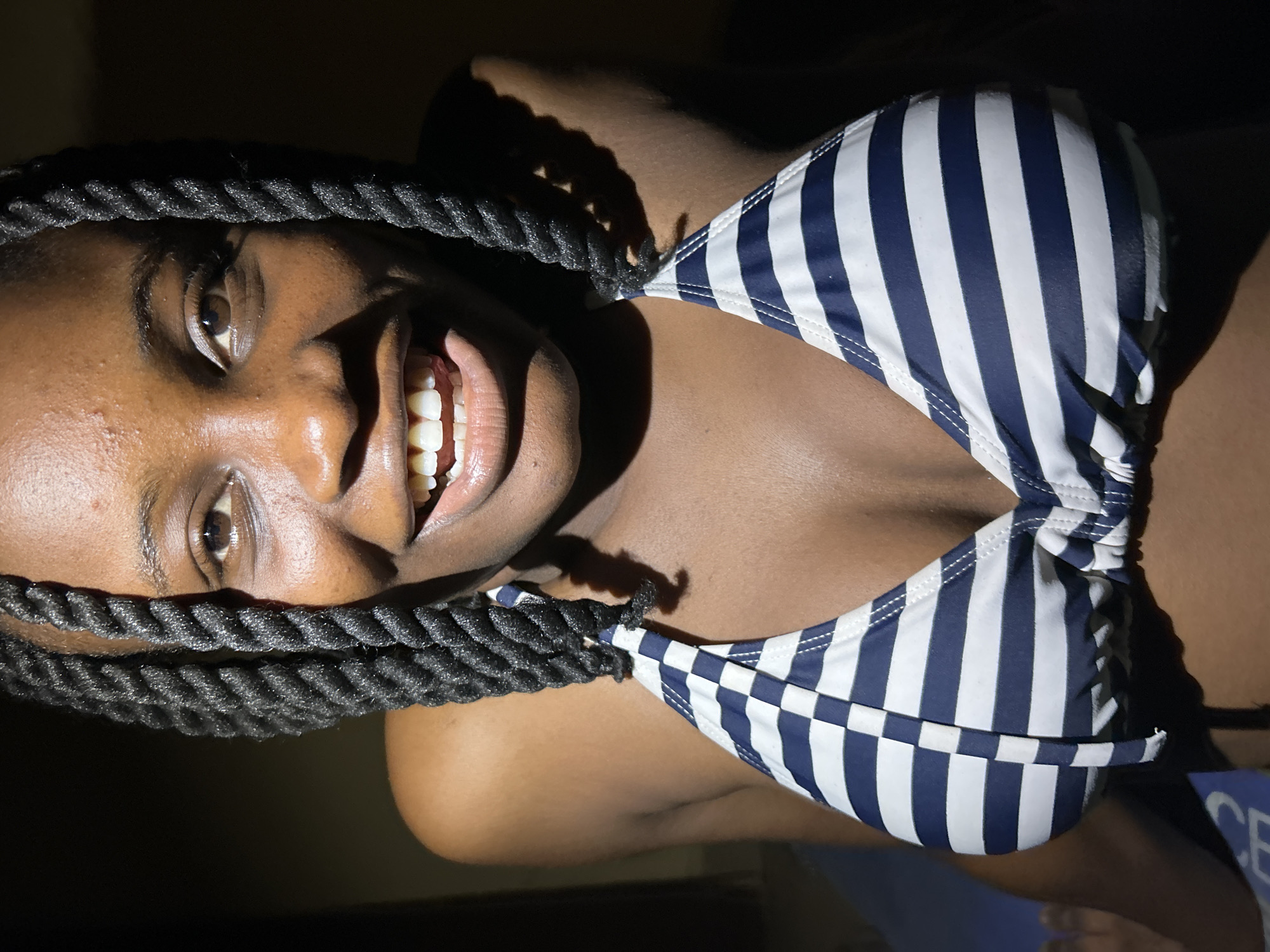 A close-up, low-angle, indoor shot shows a young Black woman with braided hair wearing a blue and white striped bikini top. She is smiling and looking directly at the camera. The lighting is dramatic, with the face well-lit and the background dark, suggesting artificial lighting in a domestic setting. The image does not contain any specific visual cues to pinpoint its location within Ihiagwa, Nigeria.