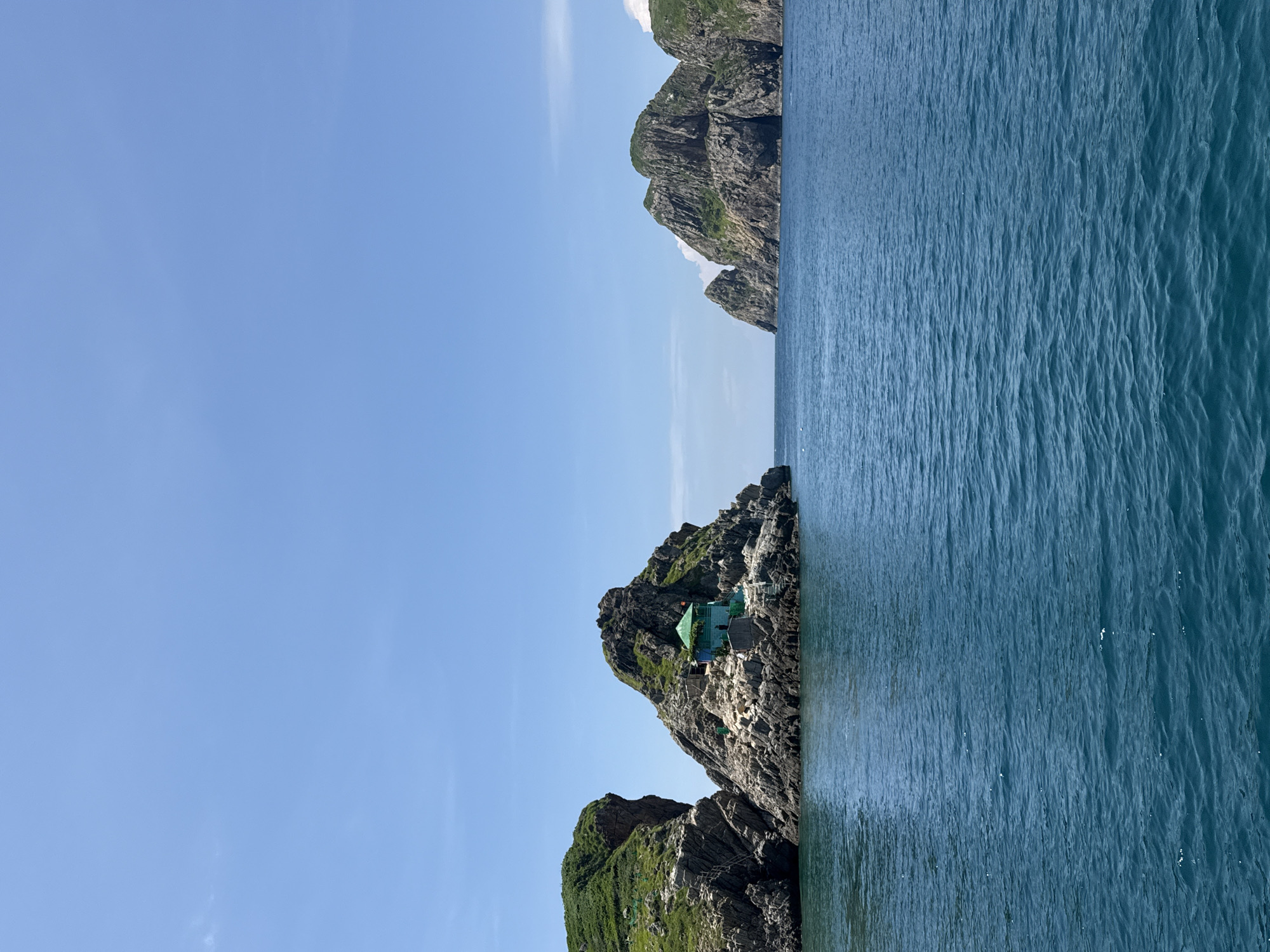 The image depicts a scenic view of islands and the ocean under a clear blue sky. In the foreground, the deep blue ocean stretches out, with gentle ripples indicating a calm sea. To the left, a cluster of rocky islands rises from the water, covered in lush green vegetation on their slopes. A small, vibrant teal-colored structure with a green roof is nestled amongst the rocks on one of these islands, accessible by a series of steps. Further to the right, more rugged, green-topped islands are visible, their rocky formations creating dramatic shapes against the sky. The scene suggests a natural, untouched coastal environment, likely a popular tourist destination due to its picturesque beauty. While the specific location is not explicitly stated as Nha Trang Ward, Vietnam, the natural landscape of islands and tropical vegetation is consistent with coastal areas in such regions.