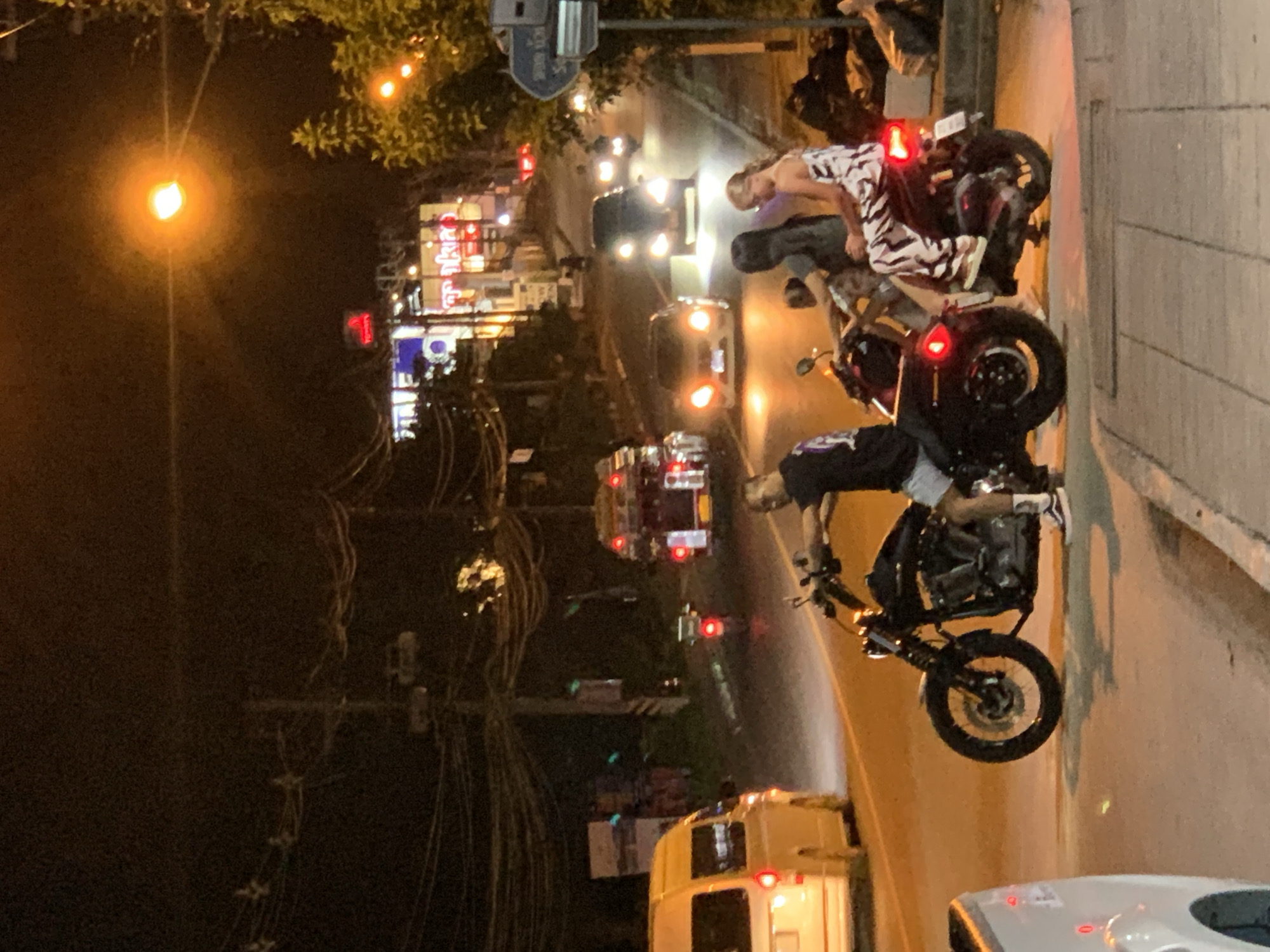 The image depicts a nighttime street scene in Baan Bo Phut, Thailand. Several vehicles are present, including a black motorcycle in the foreground carrying a man and two women. The man is on the front, driving, while the two women are seated behind him. To the left, a white van is parked. Further down the street, a red vehicle with flashing lights, possibly an emergency vehicle or a truck with decorative lights, is visible. Several cars are driving on the road, their headlights illuminated. The environment appears to be a commercial or residential area with buildings and trees lining the street. Streetlights and illuminated signs contribute to the overall lighting of the scene. The ground is paved, and sidewalks are visible. There are power lines and poles overhead. The atmosphere is that of a busy street at night.