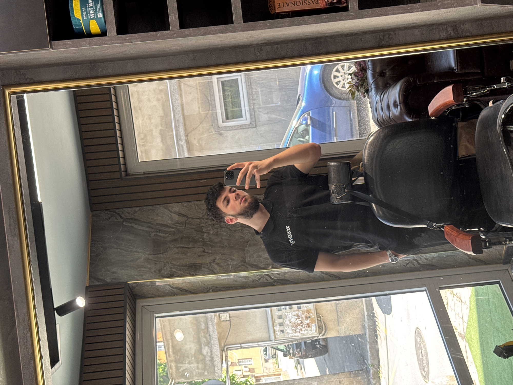 The image displays a mirror selfie taken by a young man in what appears to be a barbershop or salon. The man, with dark hair and a beard, is wearing a black t-shirt with white, stylized text (possibly "JOCAIN") and a watch on his left wrist. He holds a dark-colored smartphone to capture his reflection.
The mirror, framed in gold, reflects the interior space and the outdoor environment visible through windows. Inside, to the man's right, a dark, plush barber chair with a wooden armrest is visible. Above the mirror, shelves hold hair products, including a blue and yellow container labeled "OSWIXO MINISTYLE HAIR STYLING WAX WET WAX CLASSIC & FLEXIBLE." Another item on a shelf reads "PASSIONATE." The walls are adorned with dark, textured panels and horizontal wooden slats, with a modern wall light fixture present on the left.
Through the windows, a bright, sunny daytime scene is reflected, showing buildings and a blue car parked outside, suggesting an urban setting. The overall activity is a self-portrait taken in a professional grooming environment.