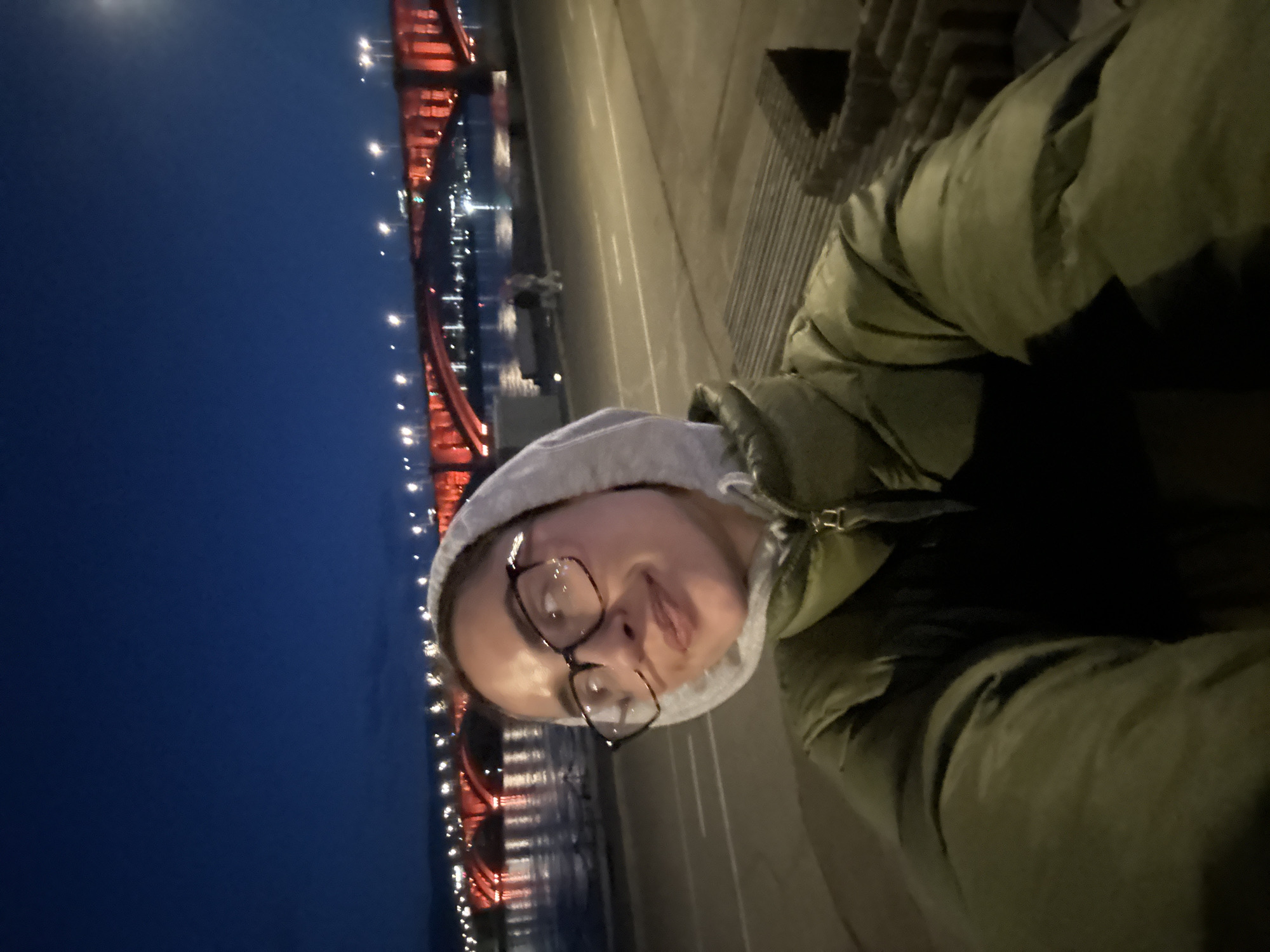 A portrait of a person with glasses wearing a grey hoodie and green jacket, with a lighted bridge visible in the background. The background indicates a night scene. Given the context of Krasnoyarsk, Russia, the bridge is likely the Vinogradovsky Bridge over the Yenisei River. The person is standing next to a wall or ledge along the riverbank.