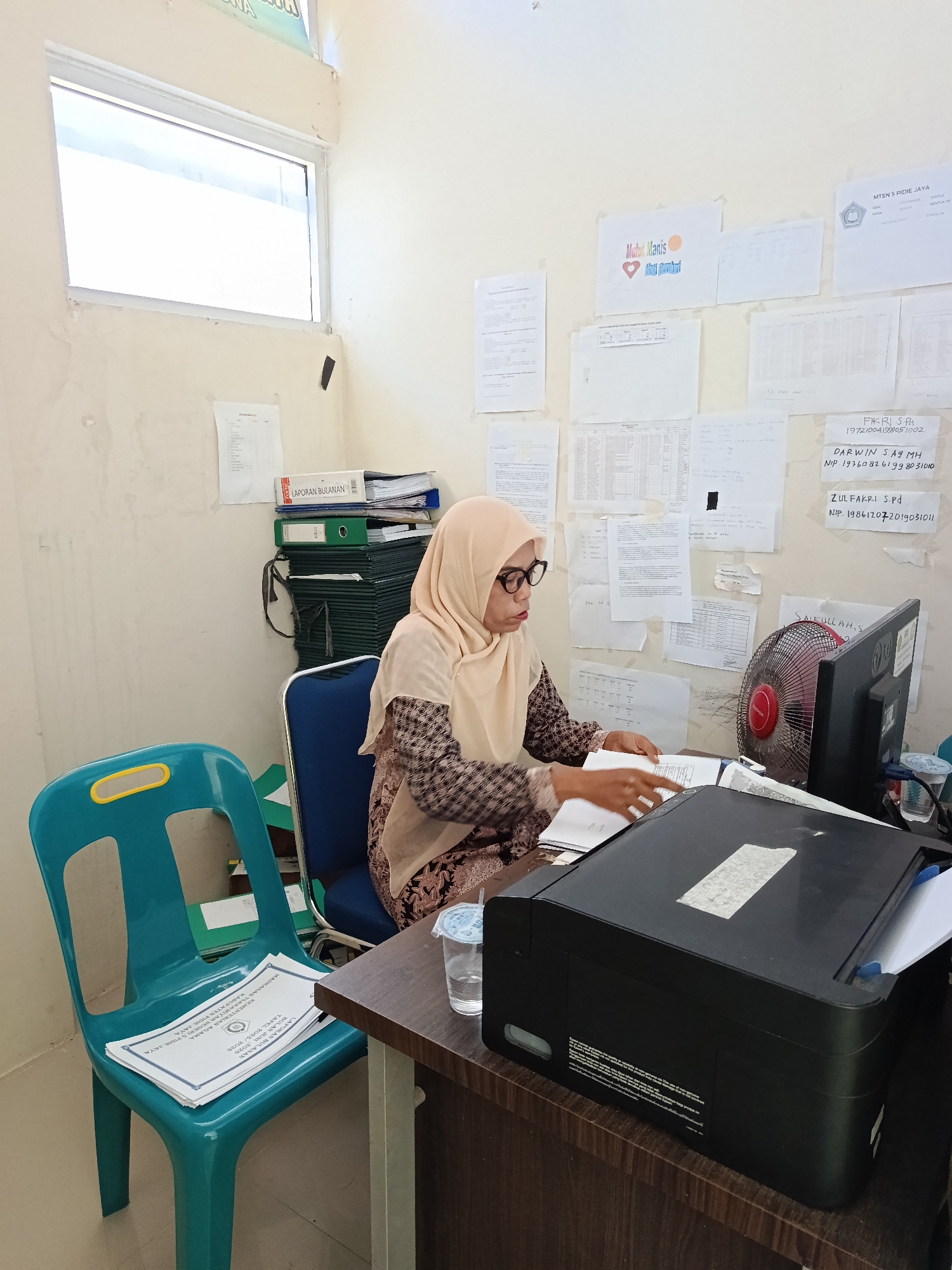 In Kuta Pungwa, Indonesia, a woman wearing glasses and a hijab is seated at a desk in a room that appears to be an office. She is looking down at documents on the desk and appears to be working. The desk is cluttered with papers, a computer monitor, a fan, a printer, and a glass of water. Behind her, the walls are covered with various documents and papers, some of which are pinned or taped up. There is a window on the left side of the room, and a stack of folders and binders is visible against the wall. A bright blue chair is situated to the left of the desk. The overall impression is that of a busy, functional workspace.