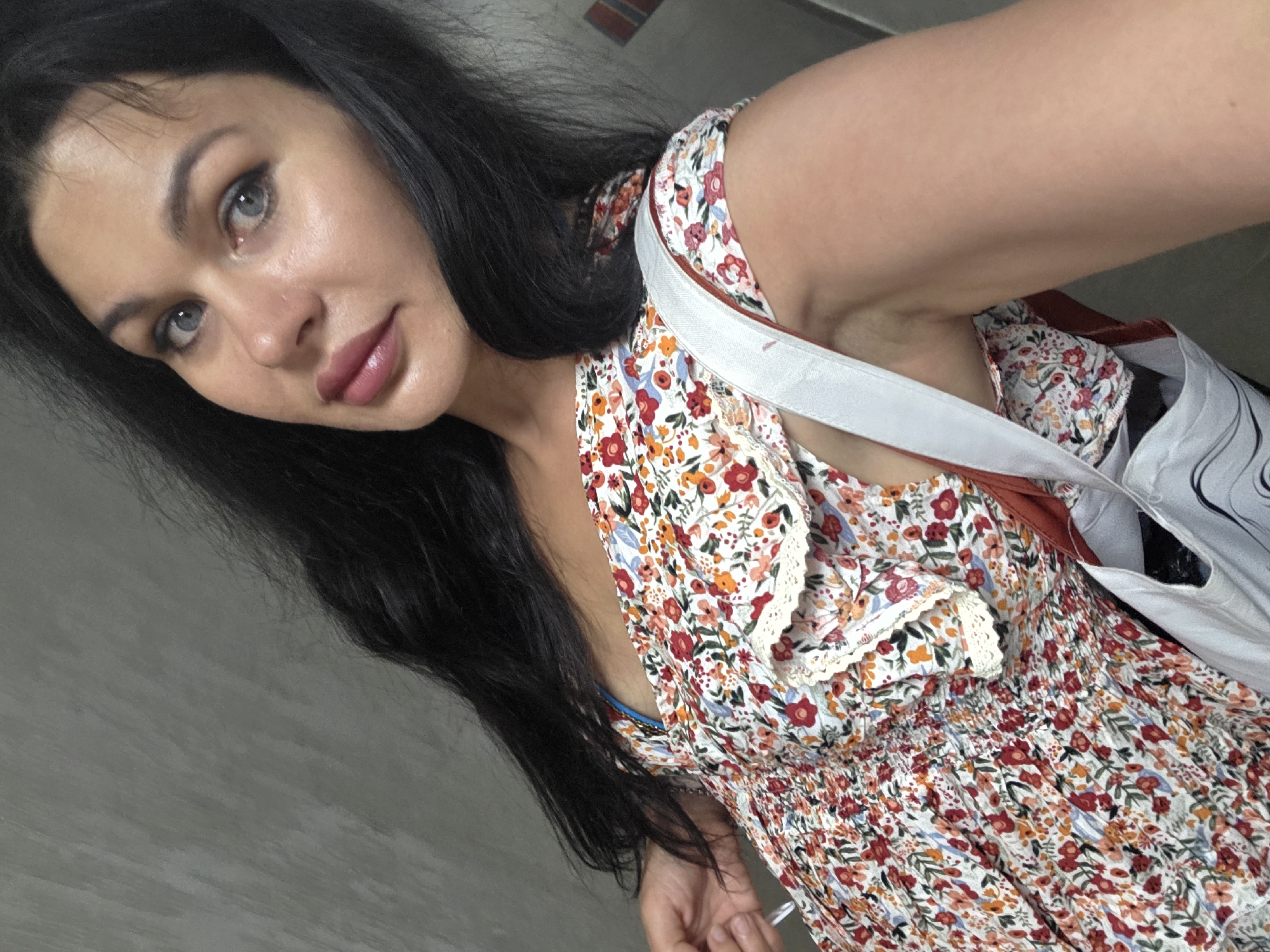 A woman is pictured indoors, against a gray, textured wall. She has long, dark hair and is wearing a floral-print dress with a white lace trim around the neckline. A white strap of a bag is visible across her shoulder. Her facial features, including her blue eyes and lips, are prominent. The lighting suggests an indoor setting, possibly a room or hallway. There is no indication of the city of Sochi, Russia, within the image itself.