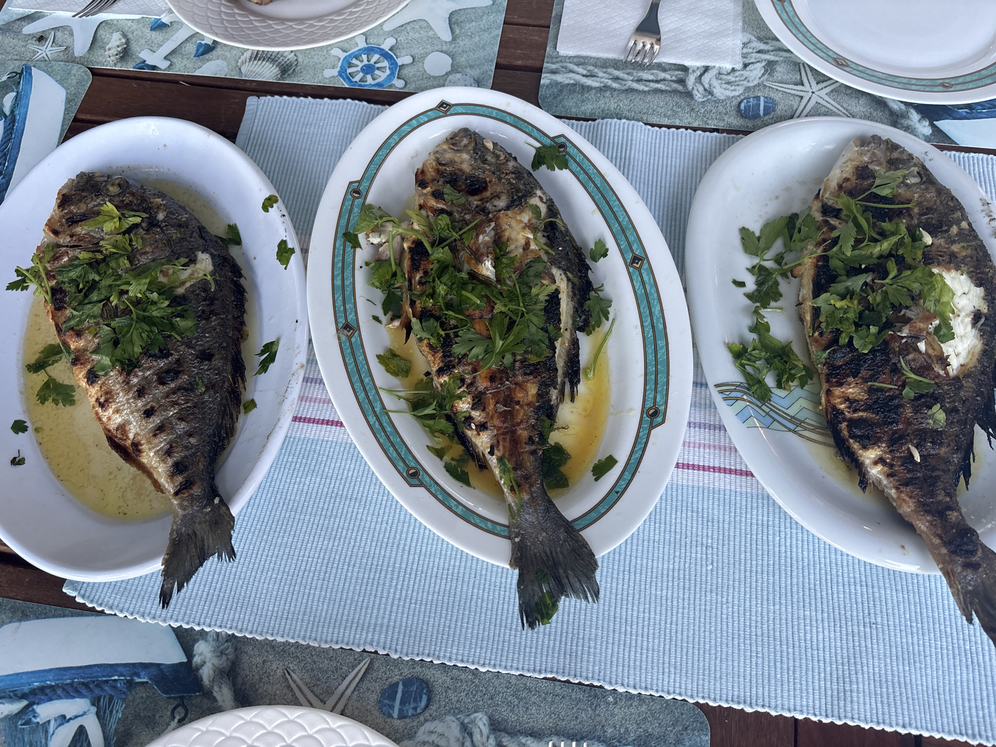 Three grilled whole fish are presented on white oval platters, garnished with chopped parsley and a light sauce. The platters are arranged on a light blue striped placemat set on a wooden table. One platter is on the left, another in the center, and the third on the right. The center fish appears to be the largest. A fork rests on a white napkin in the upper right portion of the image. The table is adorned with a nautical-themed placemat featuring anchors, ropes, and a ship's wheel. The scene suggests an outdoor dining setting, potentially in Mandra, Greece, given the context provided.