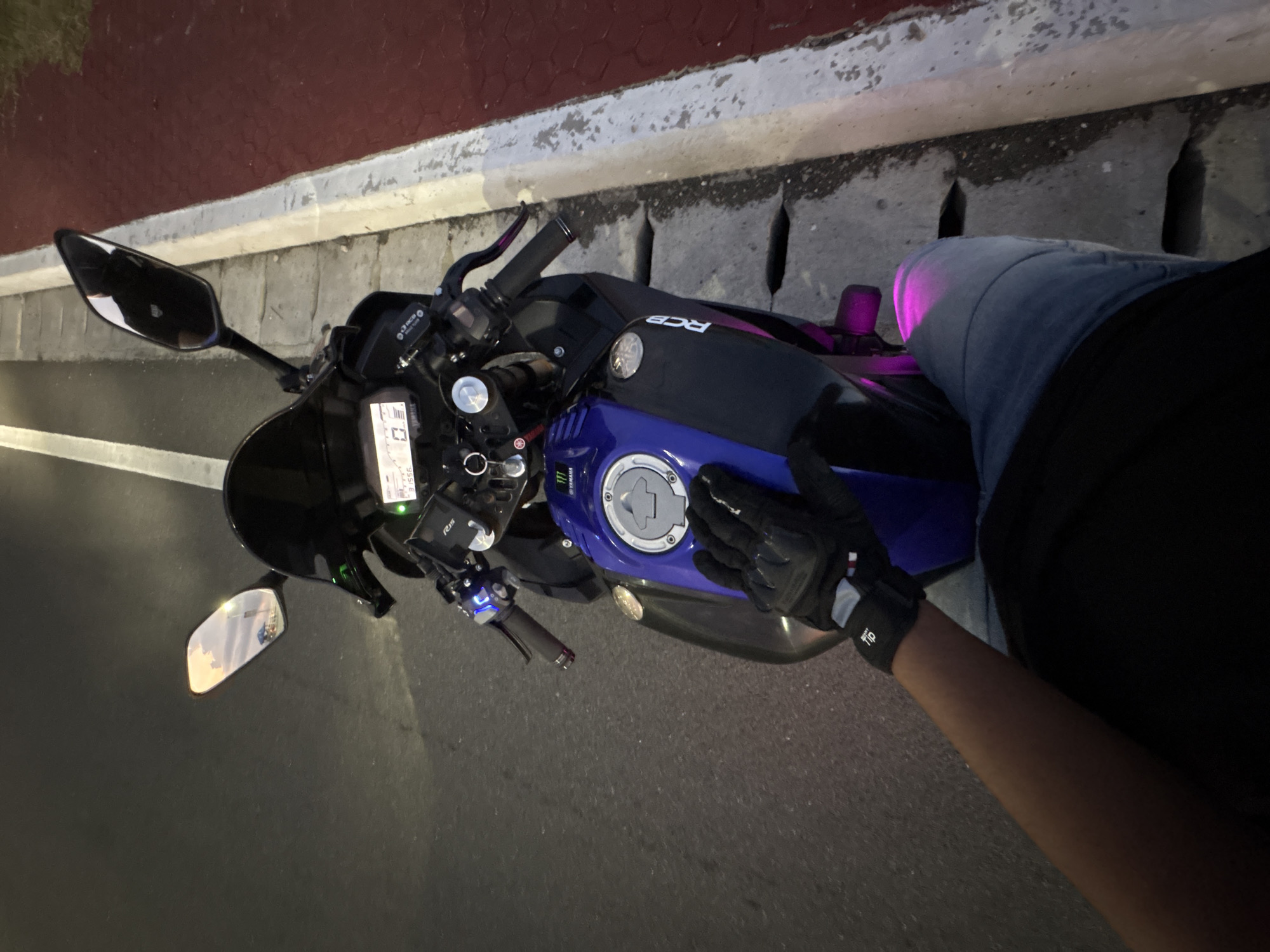 This is a first-person perspective, eye-level shot of a person's hand resting on the gas tank of a motorcycle, parked on the side of a road in Davao City, Philippines. The motorcycle is a Yamaha, identifiable by the logo on the gas tank. The rider is wearing black motorcycle gloves and blue jeans. The motorcycle has a digital dashboard displaying "0". The road appears to be paved, with a white line marking the lane. Adjacent to the road is a sidewalk or curb, and beyond that, a red tiled area, possibly a pedestrian walkway or bike lane. The scene is dimly lit, suggesting it is either dawn or dusk. The mirror of the motorcycle reflects the sky, indicating an outdoor setting. The overall impression is that of a brief stop during a motorcycle ride in an urban environment.