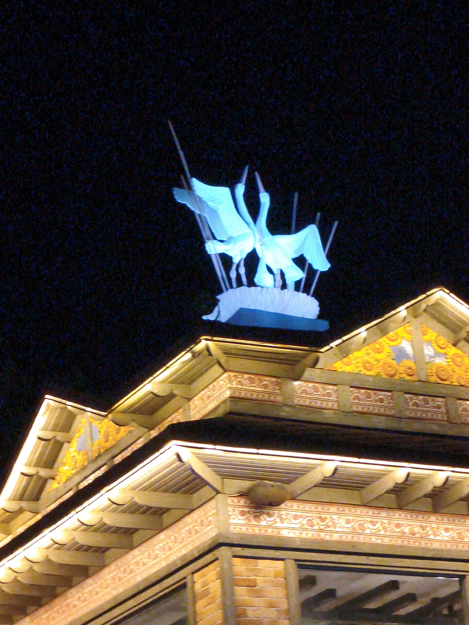 The image shows a nighttime view of a building in Divnomorskoe, Russia, with a prominent illuminated sculpture of three cranes in flight against a dark sky. The sculpture, rendered in a bright blue, is positioned above the building's roofline. The building itself features traditional architectural elements with tiled roofing and decorative banding along the eaves. The building is illuminated with warm, yellow lights, highlighting the intricate patterns and wooden beams of its construction. The overall scene suggests a festive or decorative display, likely at night.