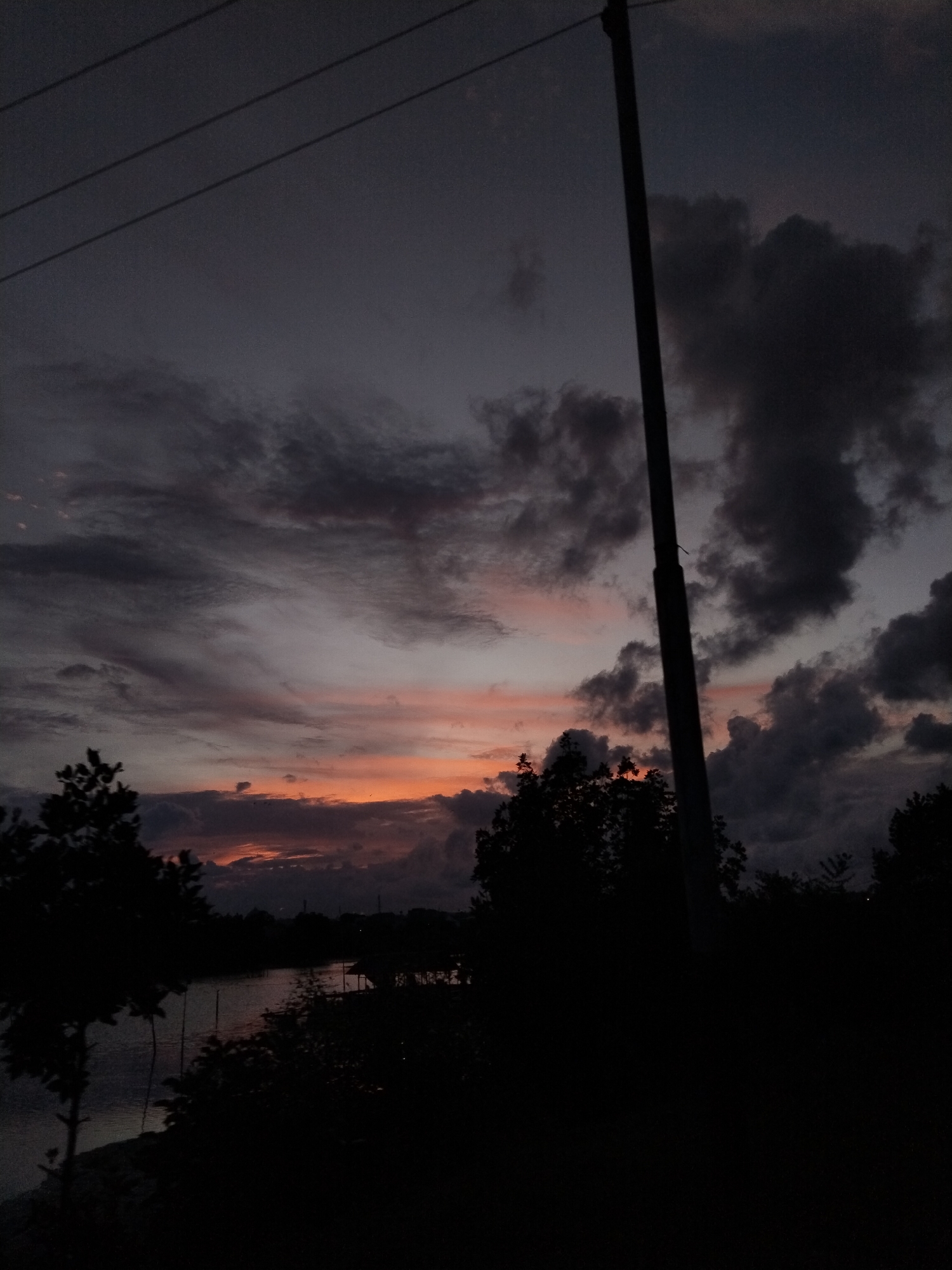 The image depicts a twilight or near-dark scene, likely at dusk, in an unspecified location in Lhokseumawe, Indonesia. The scene is dominated by a sky with dark, heavy clouds interspersed with streaks of pink and orange hues near the horizon, indicating the remnants of a sunset. Silhouetted trees and vegetation fill the lower portion of the frame, with what appears to be a body of water visible behind them. A utility pole stands prominently on the right side of the frame, with overhead power lines extending across the upper left corner. The overall scene is dim, with significant areas of shadow, making details difficult to discern. There are no discernible people or interactions visible.