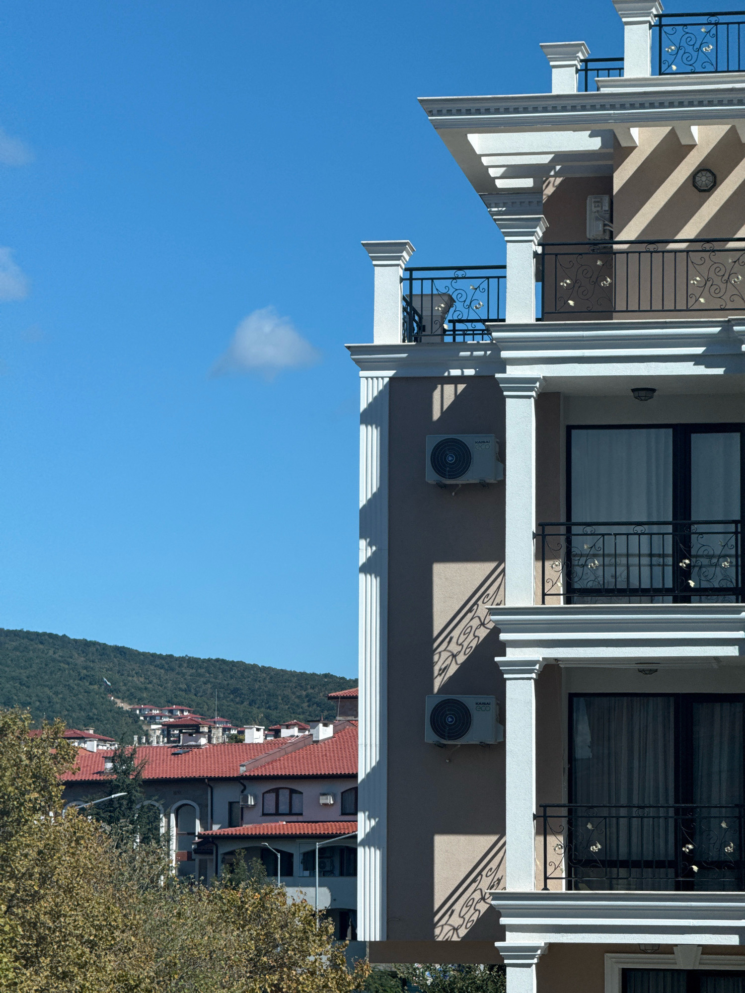 In Sveti Vlas, Bulgaria, the image captures a view of a modern apartment building with ornate white decorative elements and black metal railings. Sunlight casts diagonal shadows on the building's facade, highlighting its architectural features. Below, a cluster of traditional houses with red-tiled roofs nestles against a verdant, tree-covered hillside. The sky above is a clear, bright blue with a single small cloud. The scene conveys a sense of contemporary living integrated into a scenic, hilly landscape.