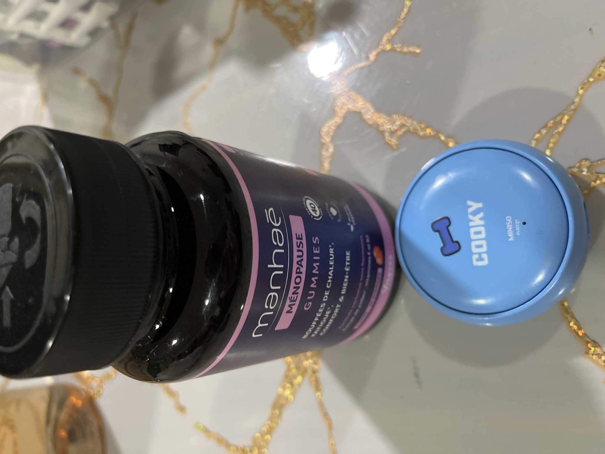 The scene, located in Unknown, Unknown, displays two primary objects resting on a light-colored, reflective surface. The surface exhibits an irregular, branching pattern of golden, glittery lines. A dark-colored, opaque plastic bottle with a black screw-on cap is situated in the upper central area. The bottle's label, featuring a purple background with pink and white text, identifies the product as "manhaé MÉNOPAUSE GUMMIES" for "BOUFFÉES DE CHALEUR, FATIGUE, CONFORT & BIEN-ÊTRE," indicating suitability from "PARTIR DE 40 ANS." A small blue circular object is positioned centrally in the lower portion of the frame. This object has a domed top displaying a blue dumbbell graphic, the word "COOKY" in white capital letters, and "MINISO BT21" in smaller text below it. The background contains blurred elements, including a warm-toned reflection to the upper left and a textured white object to the upper right.