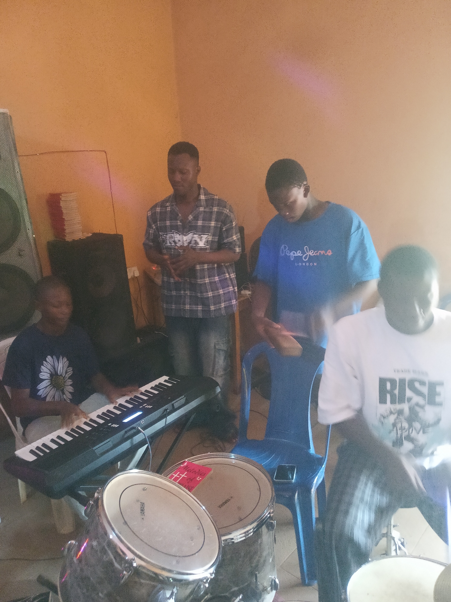 This indoor photograph from Nkwelle Ezunaka, Nigeria, depicts a band practice or jam session involving four young males. The setting is a casual room with warm, light orange-brown walls, suggesting a home studio or rehearsal space.
In the foreground and to the left, a young man wearing a dark t-shirt with a prominent white daisy pattern is seated, actively playing a black electronic keyboard. Behind him, another young man stands, dressed in a plaid shirt and dark pants, appearing to observe or participate vocally. To the center-right, a third individual in a blue t-shirt, which displays "Pepe Jeans LONDON" text, stands near a blue plastic chair, engaging with percussion, possibly holding drumsticks. On the far right, partially visible, a fourth young man wearing a white long-sleeved t-shirt with "TRADE MARK RISE" printed on it is seated and playing drums. Two tom-toms or snare drums are visible in the lower foreground, one with a red sticker featuring a cross-like symbol, and "YAMAHA" branding is faintly visible on another drumhead.
Large speakers or sound equipment are positioned on the left side of the room, and a stack of books or papers with red and white spines rests on a surface in the background. The room is brightly lit, with a noticeable lens flare and bright spots in the upper right, suggesting a strong light source, possibly natural light from a window or bright artificial lighting. The overall scene conveys a focused and engaged musical activity.