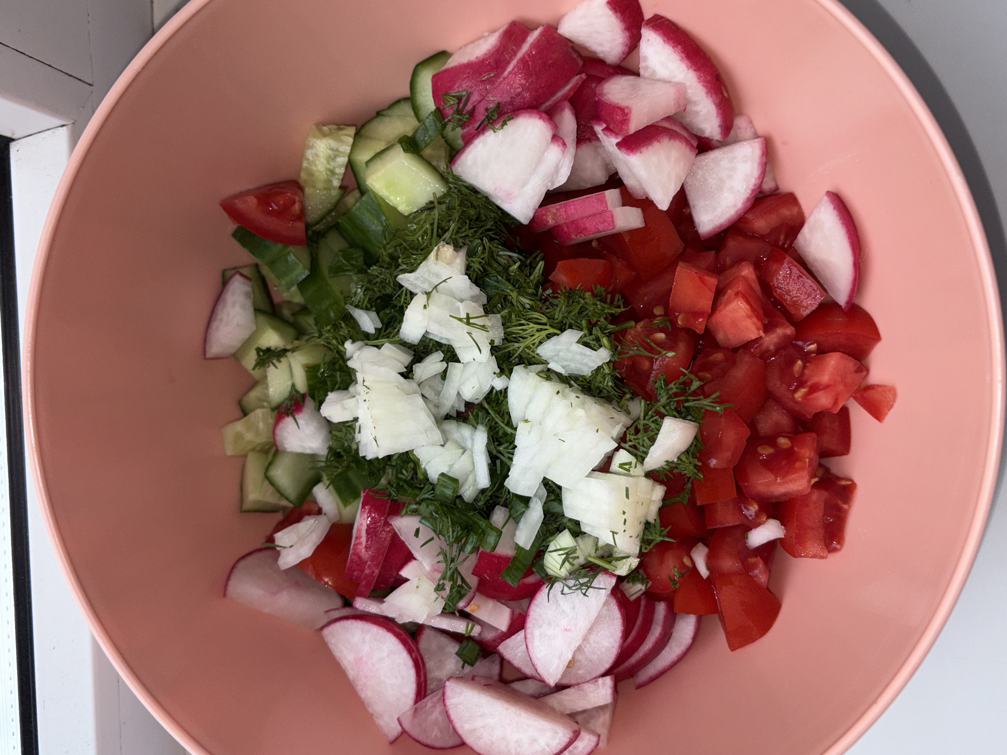 The image shows a top-down view of a pink bowl filled with a salad. The salad consists of chopped cucumbers, radishes, tomatoes, onions, and dill. The ingredients are arranged in distinct sections within the bowl. The photograph appears to have been taken indoors, with a window visible in the background. There is no information about the location of the photo, only the city of Chaikovsky, Russia is given. The image contains no people, actions, or interactions.