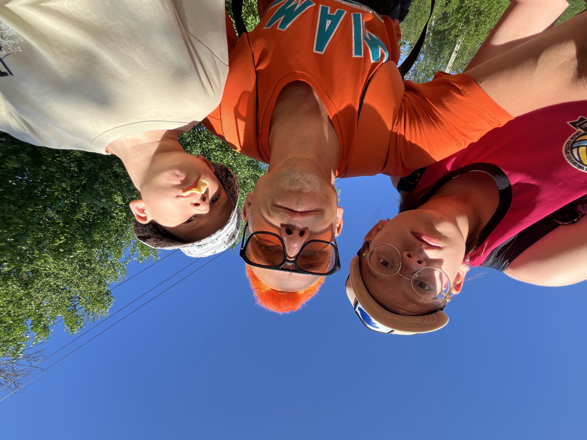 Three individuals are positioned closely together, suggesting a selfie-style photo. The person in the center has orange hair and wears large, black-framed sunglasses and an orange Miami T-shirt. To the left is another individual with a snack in their mouth and wearing a tan sweatshirt and patterned bucket hat. To the right is an individual wearing glasses and a tan and blue hat with a basketball themed top. Above them is a clear blue sky, with trees visible in the background. The location is likely Chaikovsky, Russia, as specified. The photo is taken from a low angle, with the sky dominating the background.