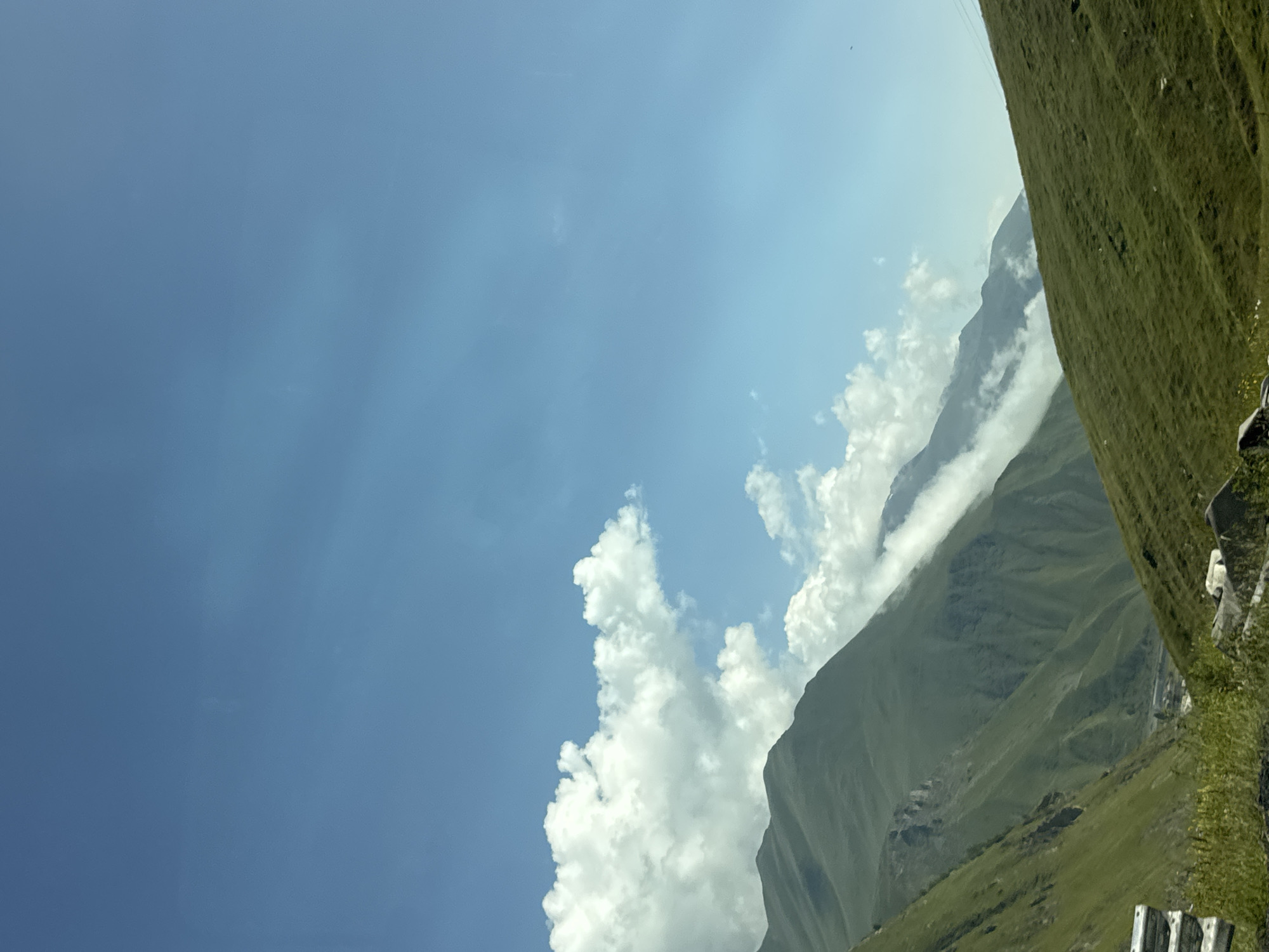 The image depicts a mountainous landscape under a clear blue sky with scattered white clouds. In the foreground, a lush green grassy slope dominates the right side of the frame. To the left, steep, verdant mountain slopes rise sharply, with some rocky outcrops visible. Wisps of clouds cling to the mid-level mountain ridges, partially obscuring them. In the distance, a valley is visible with what appears to be a small settlement or buildings nestled amongst the mountains. The overall scene suggests a rural or undeveloped mountainous region, characteristic of areas like Almasiani, Georgia, known for its natural beauty. There are no people or animals directly visible in the frame. The image appears to be taken from a slightly elevated position, possibly from a vehicle or a hiking trail.