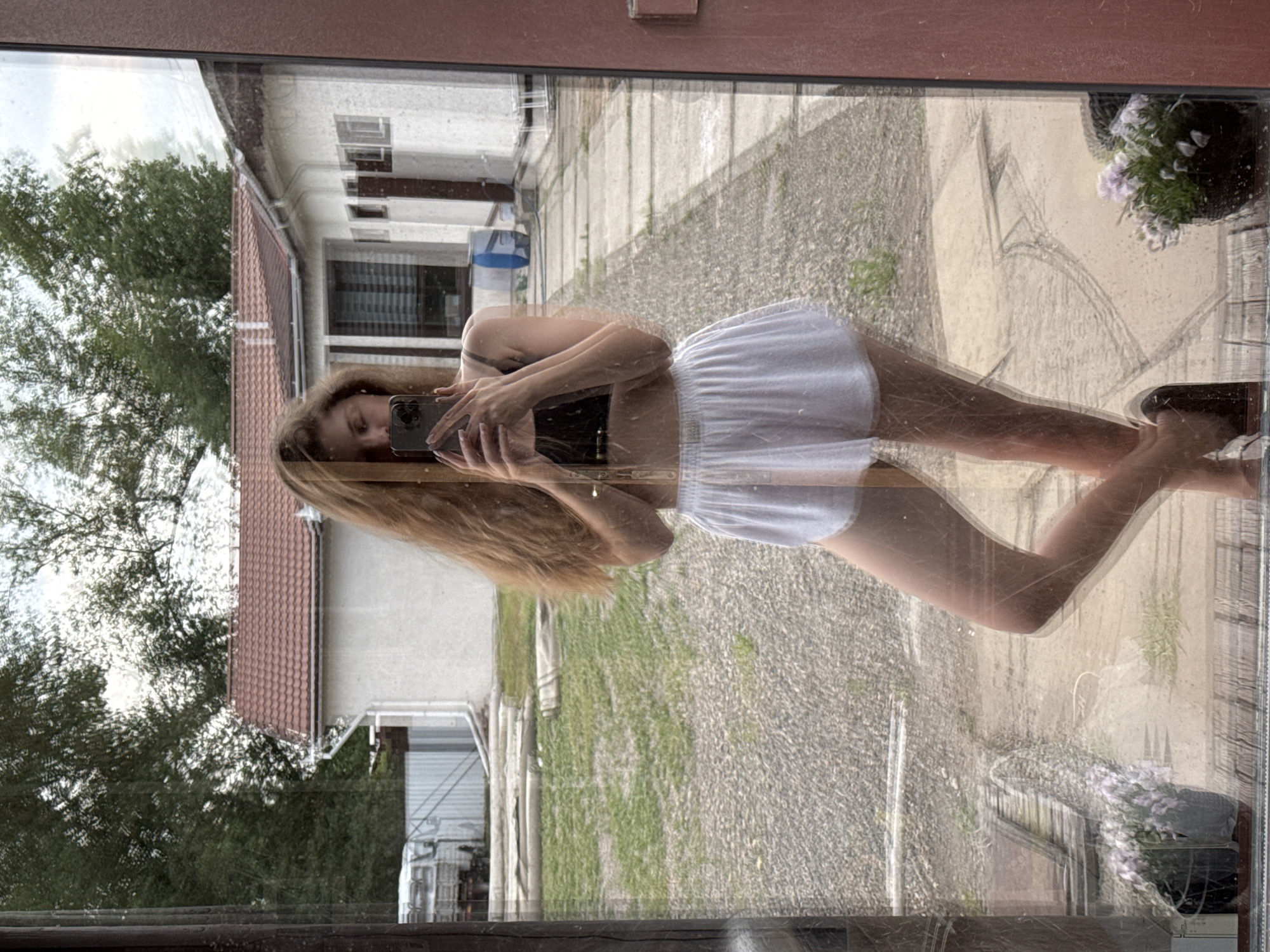 A full-body, vertical, reflected selfie of a young woman with long, blonde hair is taken from the outside of a building, looking through a window. She is wearing a black sports bra and white shorts. She is holding a smartphone in her right hand to take the picture. In the reflection, she is standing in what appears to be a backyard with a gravel path and a patch of grass. Behind her, a single-story building with a red-tiled roof is visible. In the background, trees and a white fence with a vehicle behind it can be seen. The scene is likely located in Minusinsk, Russia, given the context. The image has a slightly hazy, reflected quality due to the glass. There are two potted plants with purple flowers in the foreground of the reflection, on a wooden deck.