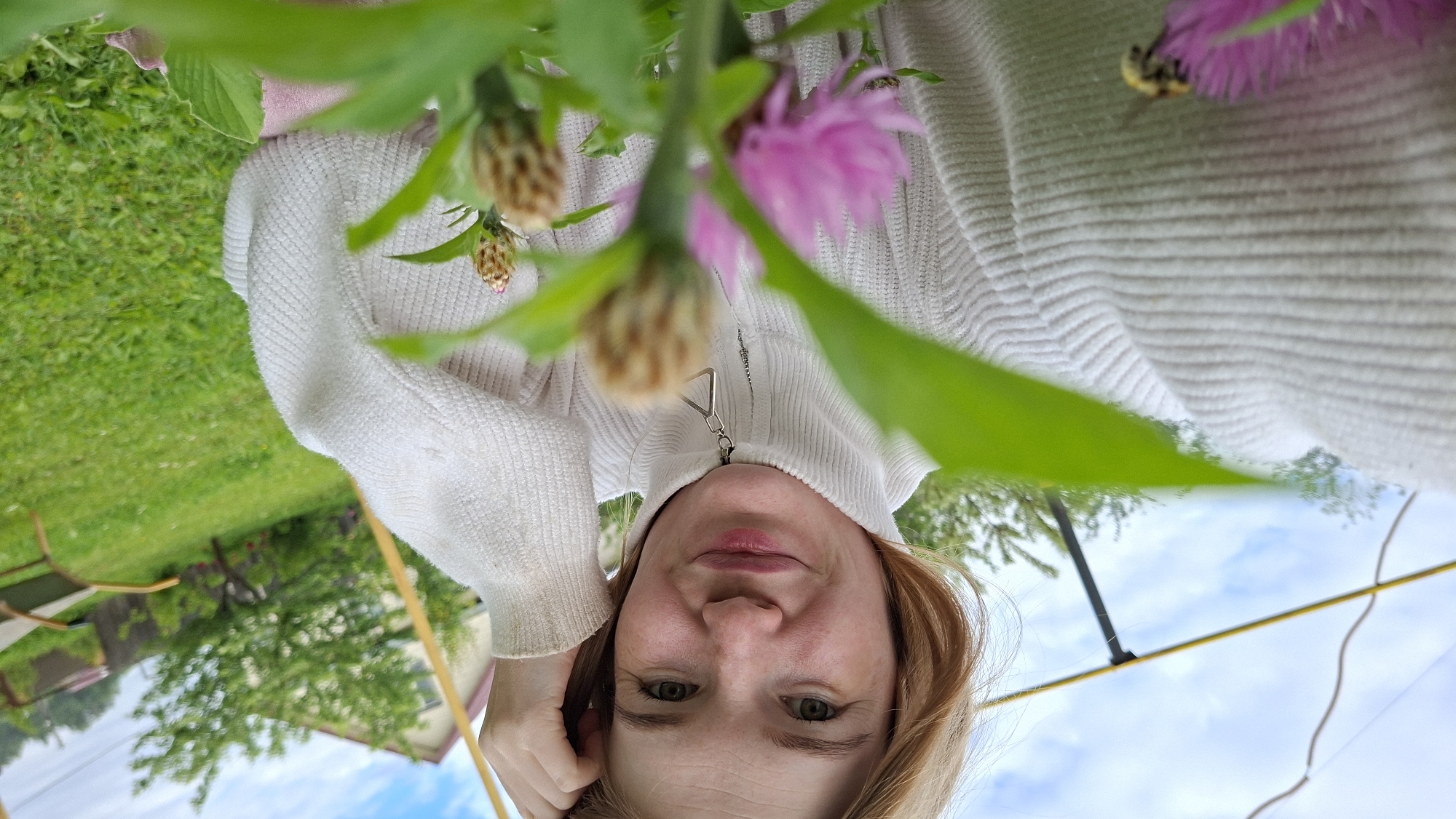 A young woman with blonde hair and green eyes is taking a selfie outdoors. She is wearing a white knitted turtleneck sweater. Her right hand is on her head, and she is smiling slightly. In the foreground, out-of-focus green leaves and pink flowers are visible, with a bee on one of the flowers. The background shows a grassy yard with trees, a house, and a fence, suggesting a residential area. The sky is partly cloudy. The location is identified as Bolszoj Bukor, Russia.