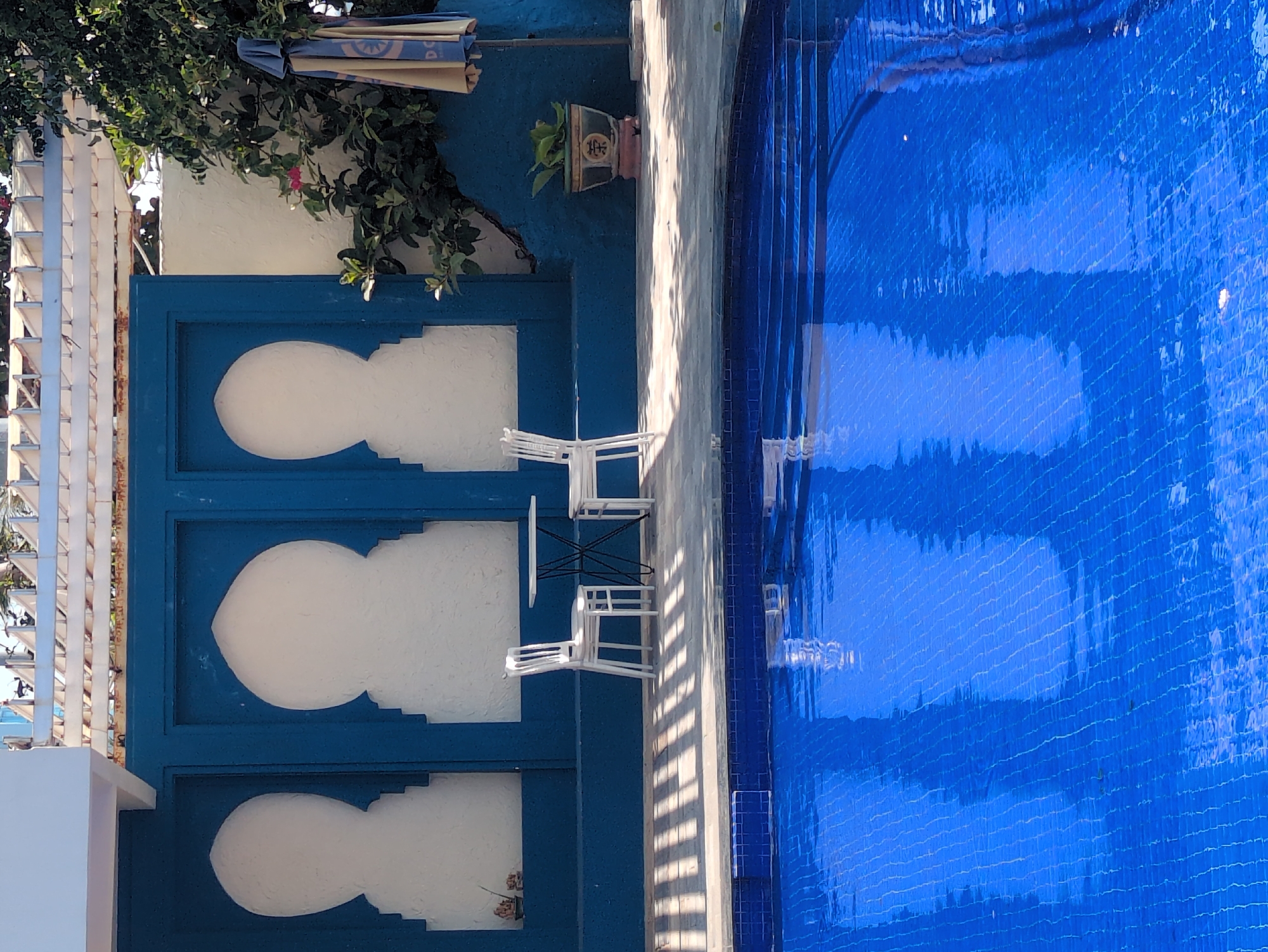The scene shows a bright blue tiled swimming pool, a wall painted in blue with white arabesque-style cutouts, and a white pergola structure covered with greenery. Two white chairs and a small black table are placed next to the wall. A potted plant hangs on the wall above the seating area. The scene may be located in South Quy Nhon Ward, Vietnam.