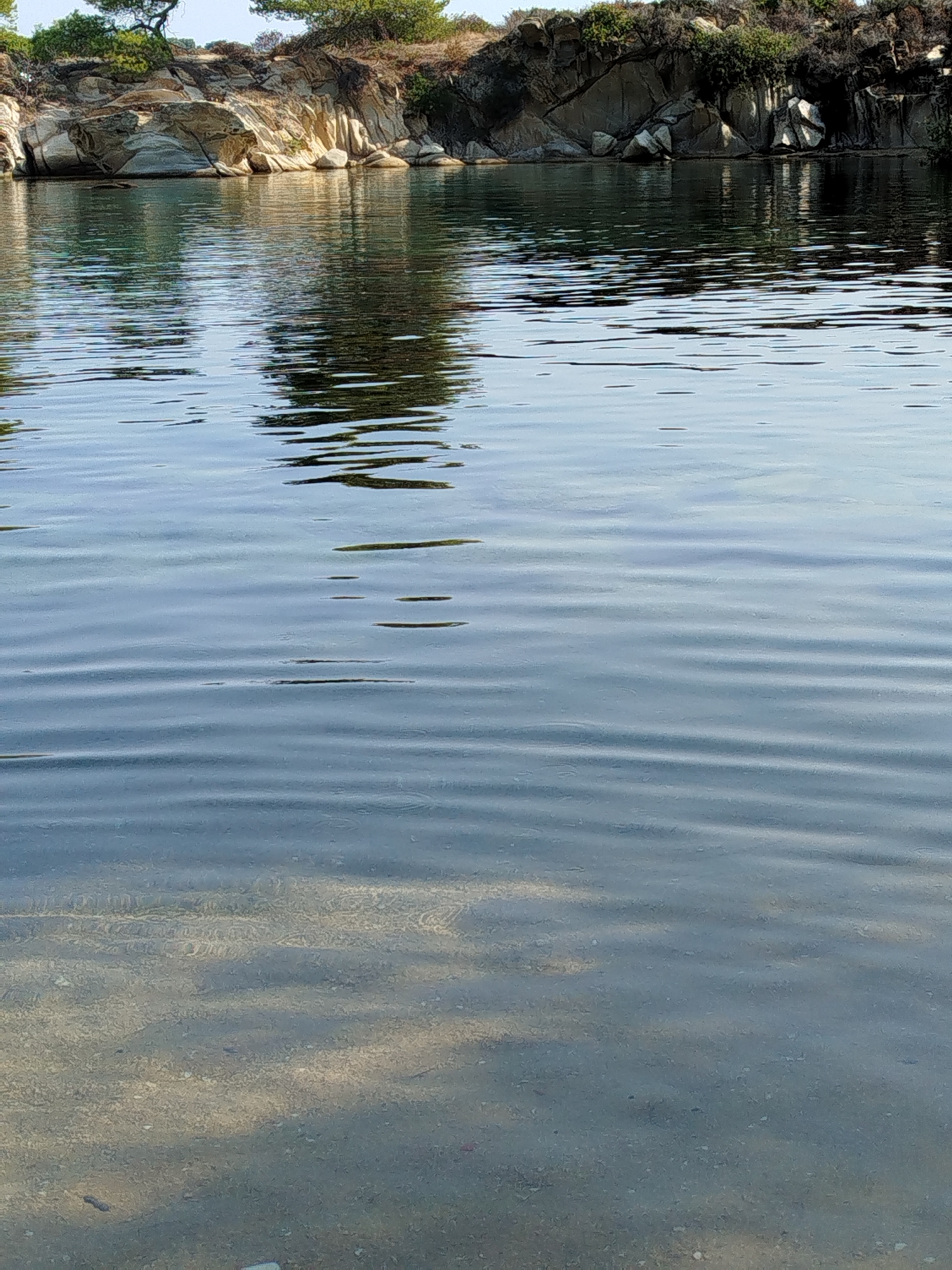 The image depicts a serene natural scene with a body of water in the foreground and a rocky coastline with sparse vegetation in the background. The water exhibits ripples, suggesting a gentle movement or breeze. Sunlight reflects off the water's surface, creating shimmering patterns. The seabed is visible in some areas, showing a sandy bottom with scattered pebbles and rocks. The coastline is characterized by rugged, layered rock formations and greenery, indicative of a Mediterranean landscape. The overall impression is one of tranquility and natural beauty. This scene is located in Sithonia, a municipal unit within the Chalkidiki regional unit of Greece.