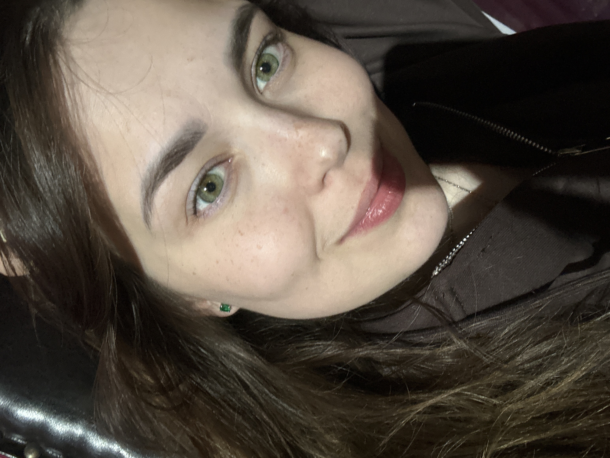 A young woman with light skin, freckles, and green eyes poses for a selfie. She has long, dark brown hair that is slightly messy. She is wearing a dark brown zip-up hoodie and a small, emerald green stud earring in her left ear. Her lips are slightly parted and appear to be wearing lip gloss. The background is dark and out of focus, with some shiny surface visible on the left. The image was taken indoors. There is no information available regarding the location in the city of Zhebrei, Russia.