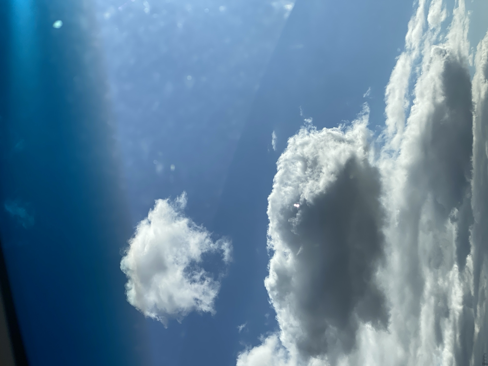 A photograph taken from below looking up at a bright blue sky filled with clouds. There are several white and gray clouds scattered across the sky, with some appearing fluffy and others more dense and shadowed. A faint horizontal line near the top of the image suggests a boundary or obstruction, possibly a window or frame. The lighting indicates a sunny day. The image does not contain any people, buildings, or recognizable landmarks of Heringen/Helme, Germany.