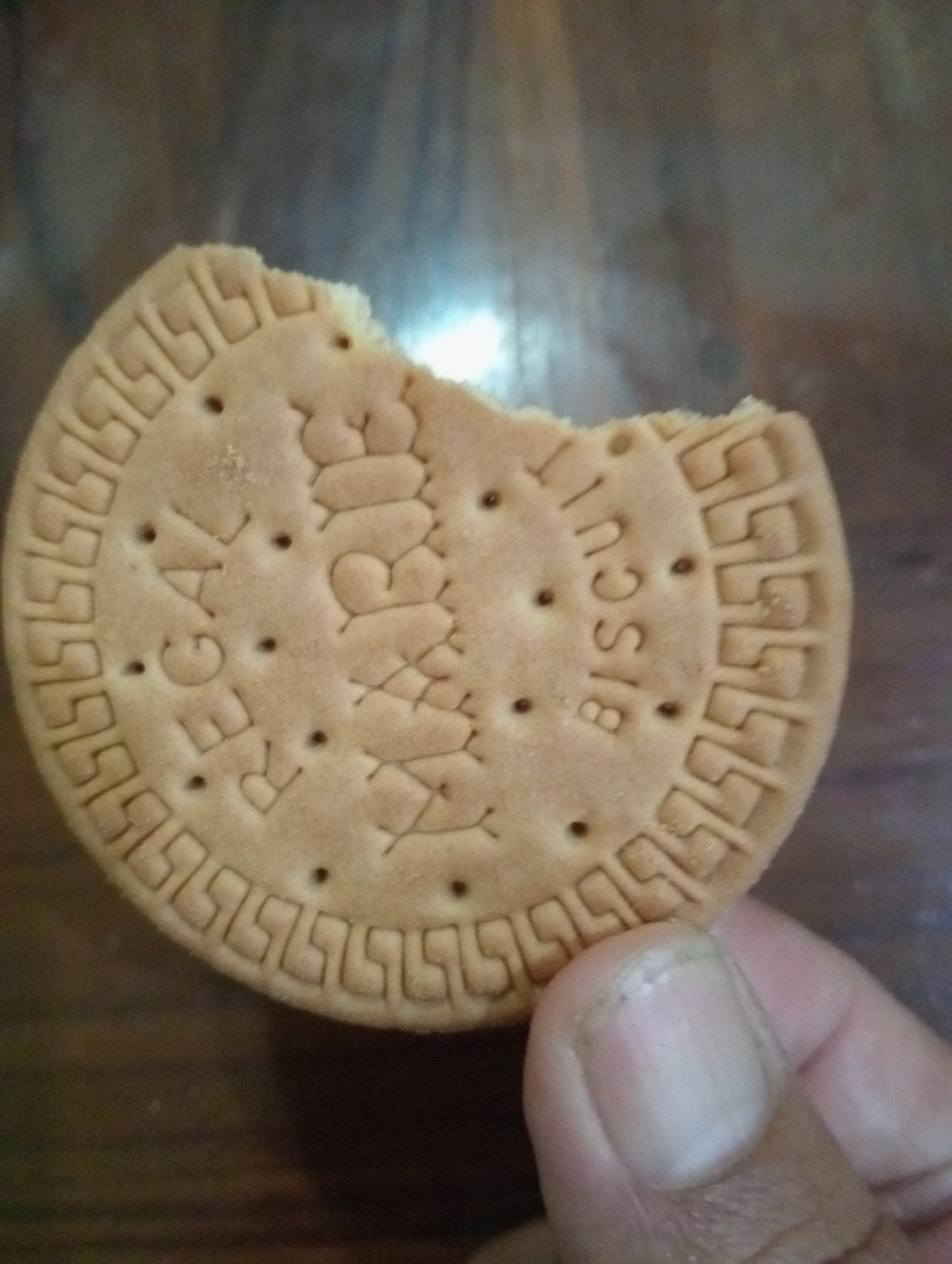 A close-up, high-angle shot captures a partially eaten, circular biscuit with a decorative embossed pattern and text. The biscuit is light brown and sits on a dark brown, wooden surface. The text embossed on the biscuit reads "REGAL BISCUIT". A human finger, with clean fingernails, is visible in the lower right corner, appearing to hold the biscuit. The background is blurred, showing a dark, textured surface. The image was taken in Sidoharjo, Indonesia.