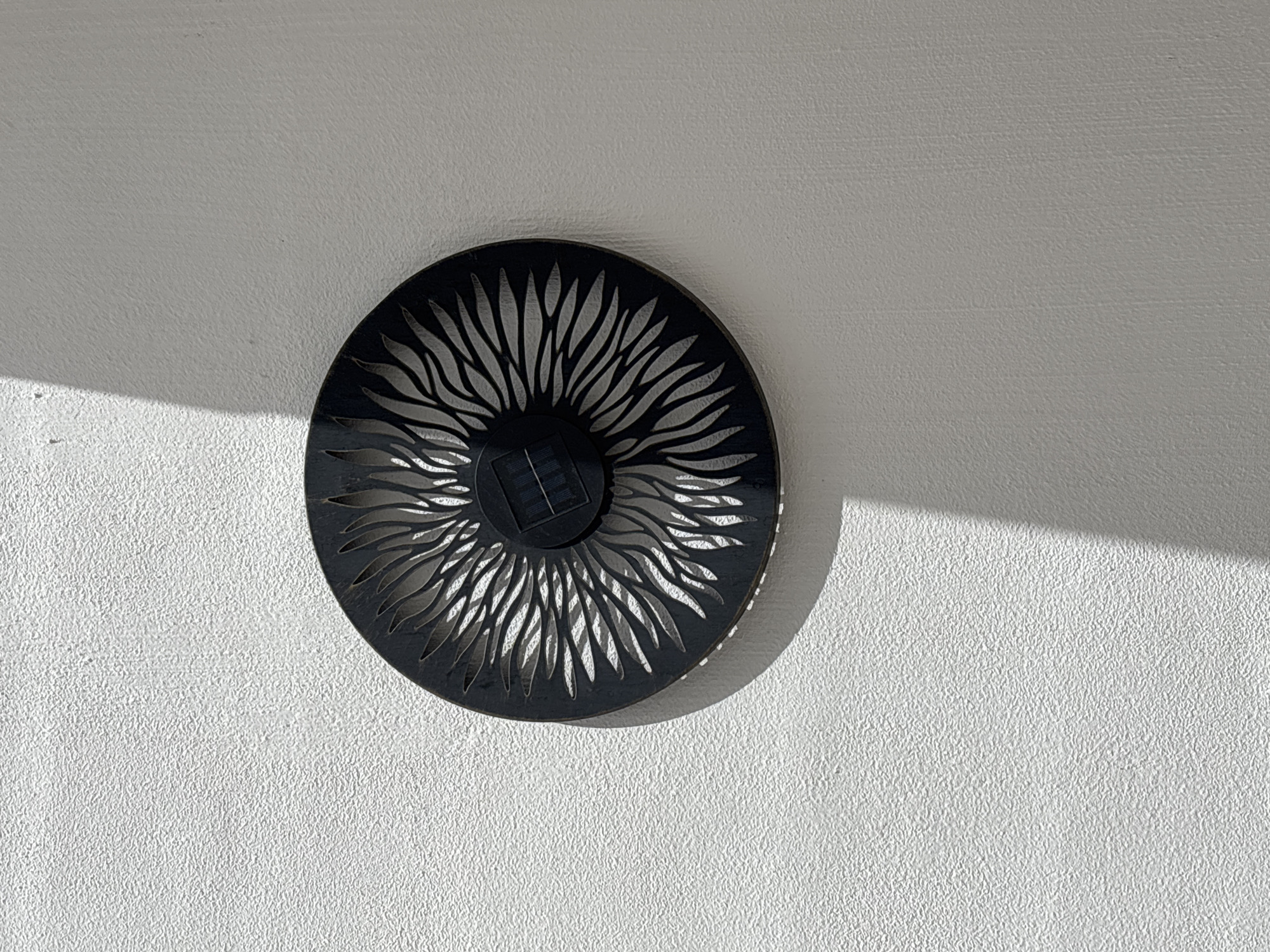 A black solar-powered light fixture with an ornate, sunburst-like design is mounted on a textured, white wall. The fixture is circular and features a central square solar panel. Light casts a distinct shadow across the wall to the left of the fixture. The setting appears to be an outdoor environment, possibly a wall in the city of Orihuela, Spain, given the context of the query. No people are present in the image.