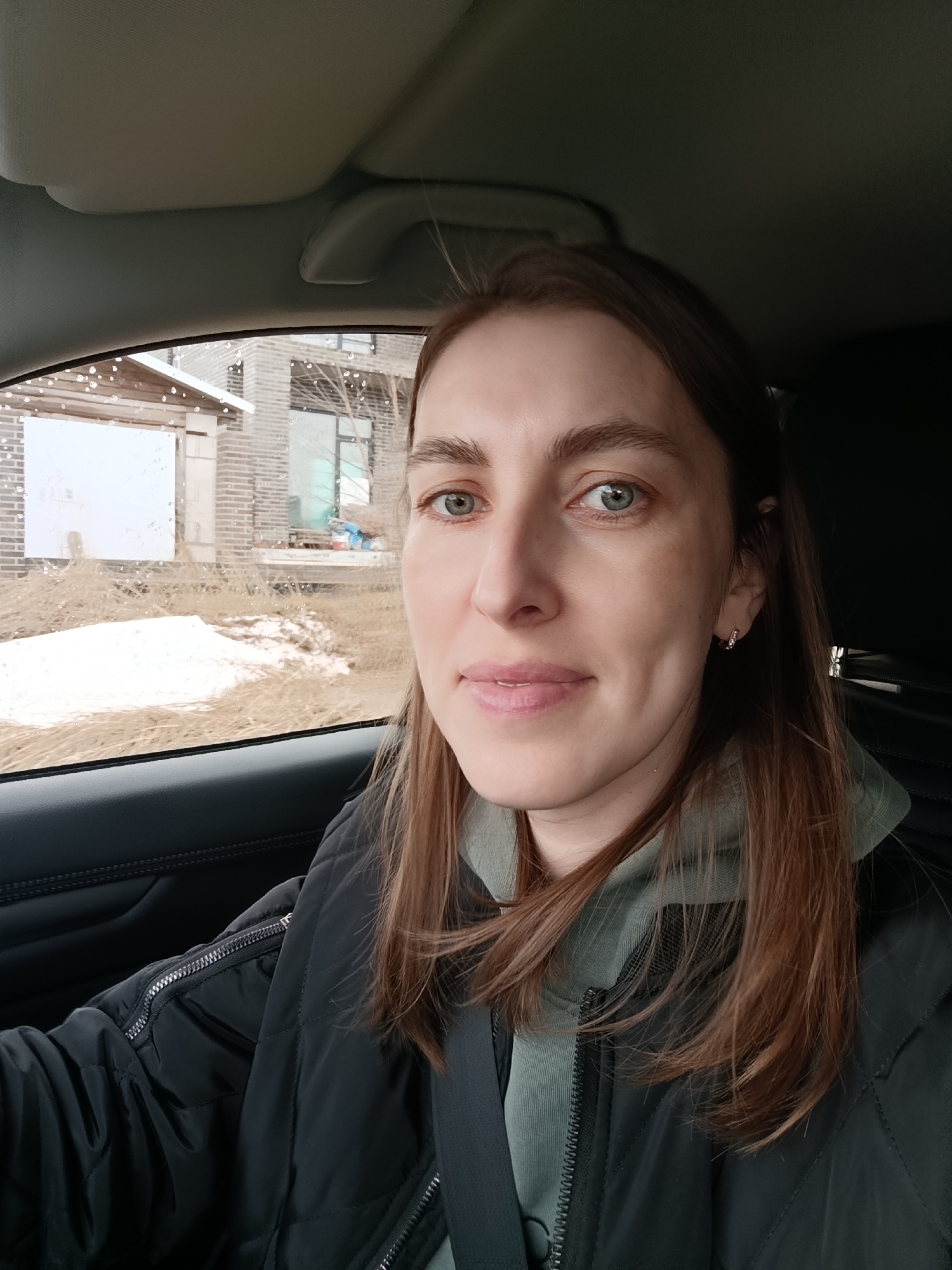 A woman with light brown hair and green eyes is seated in a car. She is wearing a black quilted jacket and a green hooded sweatshirt. A seatbelt is visible across her chest. Through the car window, a building with brick accents and a patch of snow-covered ground with dry grass are visible. The setting appears to be in a residential area, possibly in Chaikovsky, Russia. Rain or droplets are apparent on the car window.