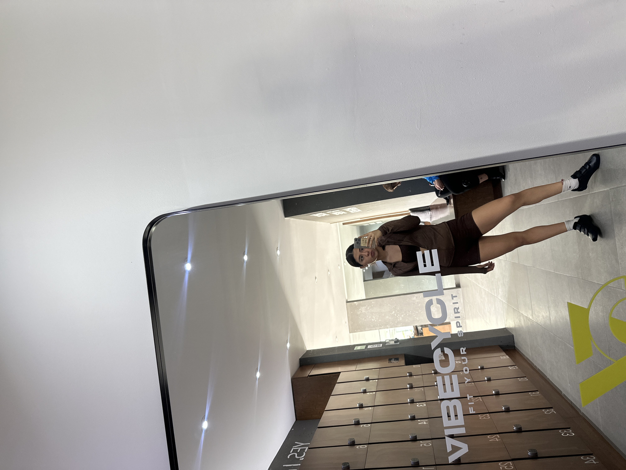 A woman is reflected in a full-length mirror in a gym locker room in San Bernardino Tlaxcalancingo, Mexico. She is wearing brown athletic wear, including a long-sleeved top and shorts, and black sneakers with white socks. She holds a phone, presumably taking the picture. Lockers line the lower half of the mirror's reflection, and gym signage is visible on the mirror and the floor. Another person is partially visible in the background. The room is brightly lit.