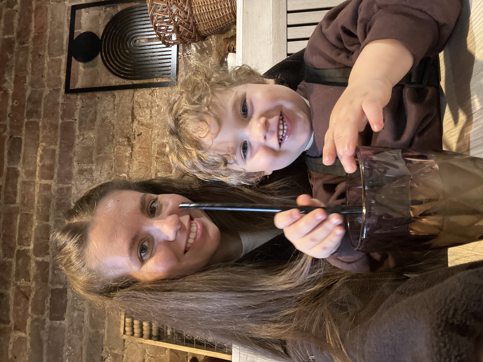 A woman and a toddler are posing for a photo in an indoor setting. The woman has long brown hair and is holding a black straw. The toddler, with curly blonde hair, is sitting in what appears to be a high chair and is reaching for a glass with a dark liquid. The background includes a brick wall with a framed decorative art piece, a wicker basket hanging above, and part of a shelf or cabinet. It is not possible to verify the location is the city of Klin, Russia, from this image alone.
