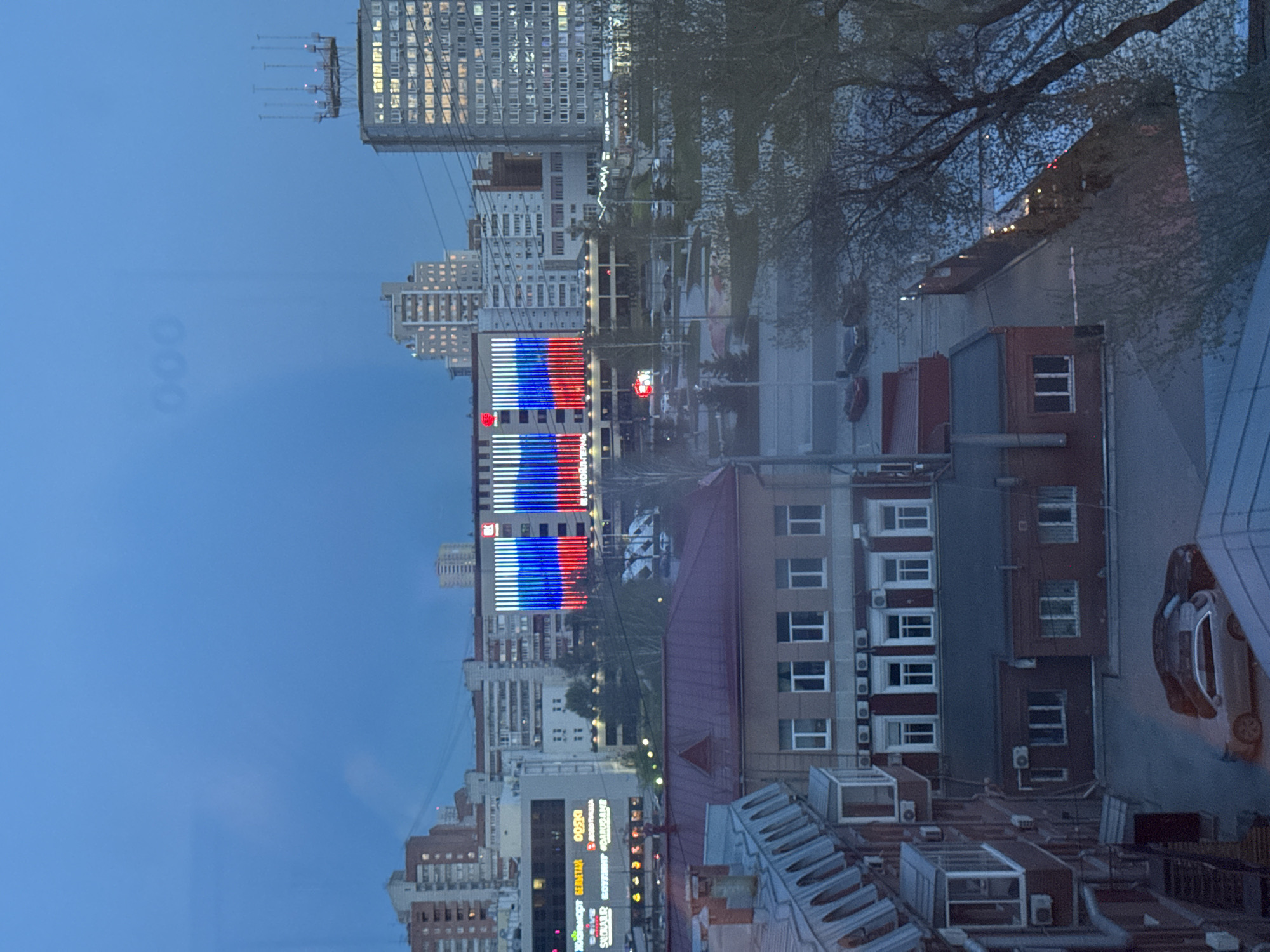 This image shows a cityscape at dusk in Perm, Russia. A building is illuminated with the colors of the Russian flag (white, blue, red). Other buildings, some residential and some commercial, are visible around it. A street and a few cars can be seen. The view appears to be from a high vantage point, possibly from a window or a balcony. The presence of a car near the bottom-right of the image suggests that the viewer is on a roof or other elevated structure.