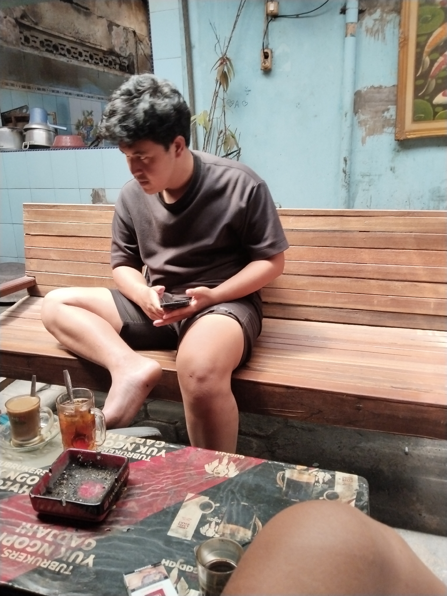 A young man is seated on a wooden bench in an outdoor setting. He is wearing a dark grey t-shirt and dark shorts, and is holding a smartphone in his hands. To his left, on a table in front of him, are two drinks in clear glasses and an ashtray filled with cigarette butts. The table's surface is decorated with a red and black graphic pattern. The background shows a blue-painted wall with some peeling paint and a framed artwork of koi fish. A plant with thin leaves is growing near the wall. The overall environment appears to be casual and possibly a roadside cafe or a backyard gathering spot in Kecamatan Pati, Indonesia.