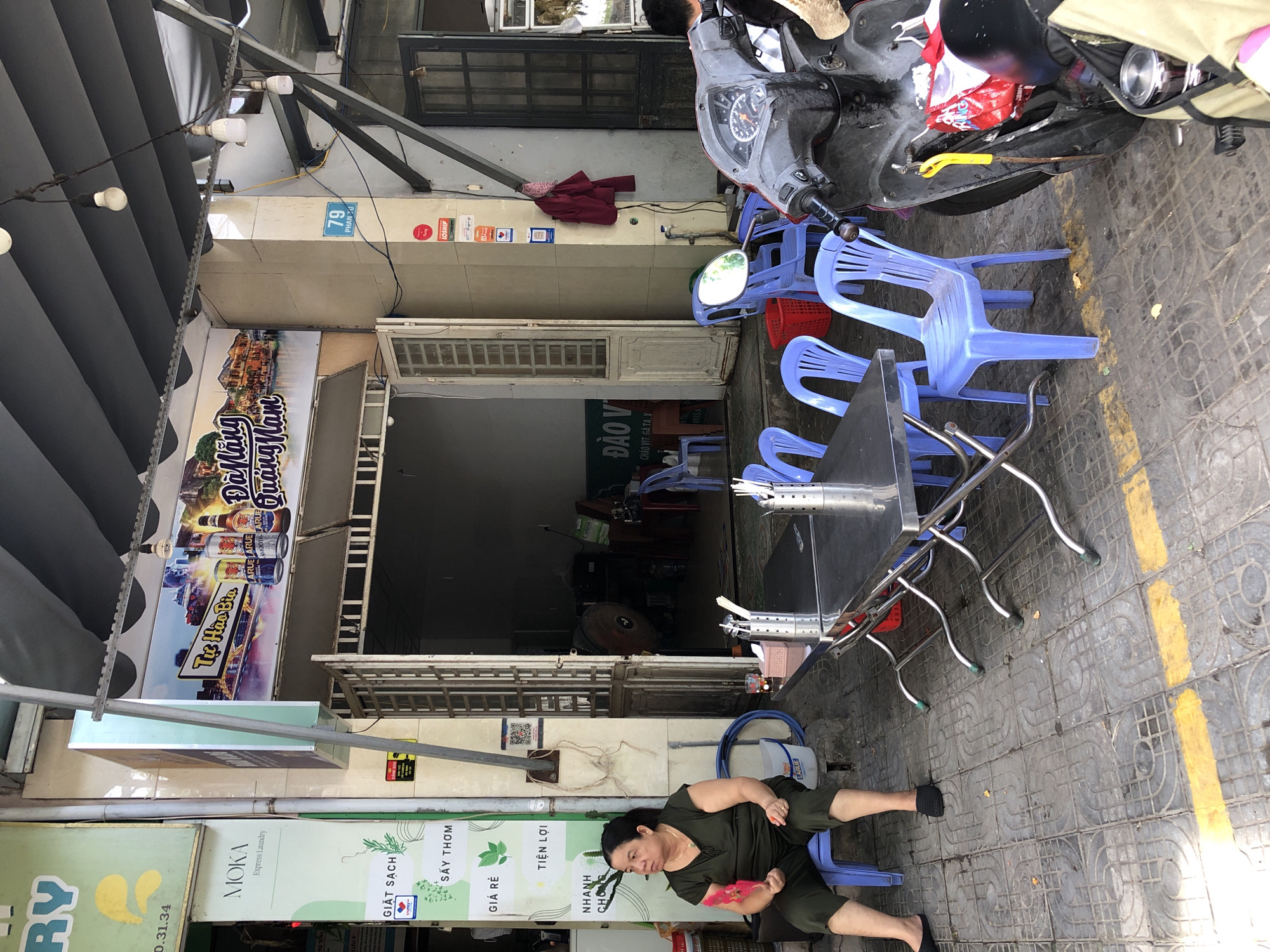 The image depicts a street scene in Đà Nẵng, Vietnam, with an outdoor seating area for a business. A woman sits on a blue plastic chair to the left, holding a red fan. In front of her and extending to the right is a metal folding table with several blue plastic chairs arranged around it. There are metal containers with cutlery on the table. A black scooter is parked to the right of the chairs. The background shows the entrance to a building with an open door revealing interior elements such as a fan. Signage is visible above the entrance, with Vietnamese text and images of beer bottles and buildings. Additional signage with Korean text and branding such as "MOKA Express Laundry" is present on the left side of the image. The ground is paved with square tiles, and a yellow line is painted on the pavement. The overall impression is of a casual, open-air establishment.