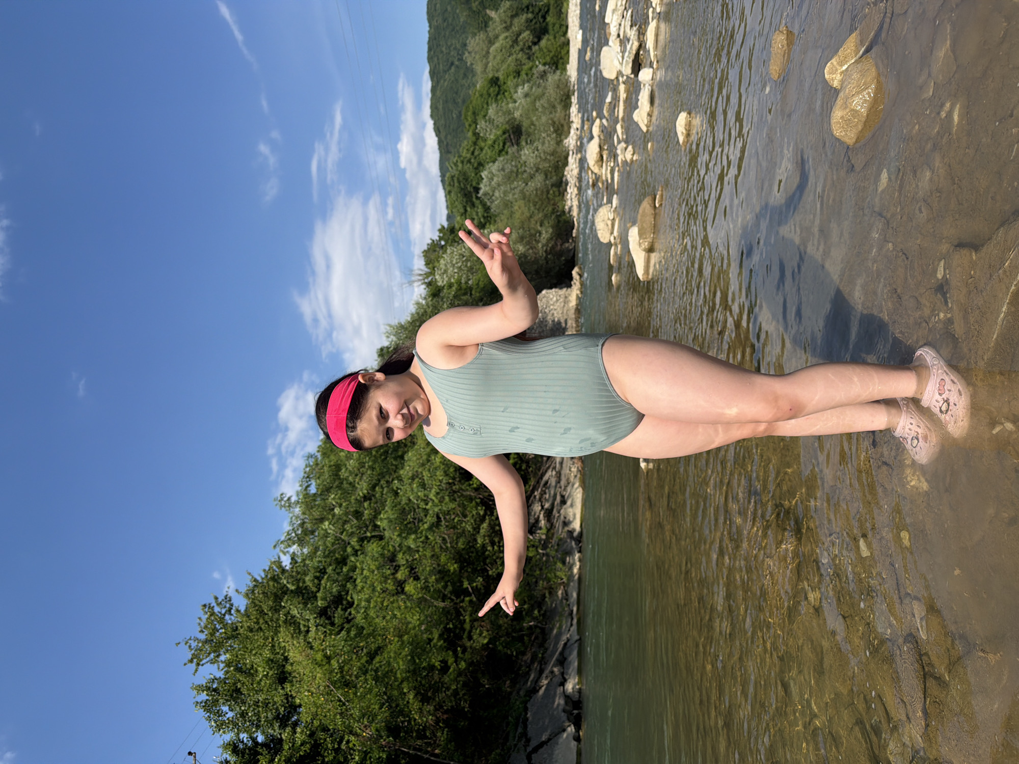 A young girl in a mint green swimsuit and a pink headband is standing in a shallow, rocky river. She is making a peace sign with her left hand and pointing down with her right hand. Her legs are submerged in the clear water, and she is wearing pink shoes with a white pattern. The riverbed is visible, covered with smooth stones and some larger rocks. In the background, lush green trees and hills are visible under a bright blue sky with scattered white clouds. The sun appears to be high in the sky, indicating it is likely daytime. The overall mood seems to be playful and relaxed, suggesting the girl is enjoying her time outdoors. The location is identified as Krasnoye, Russia.