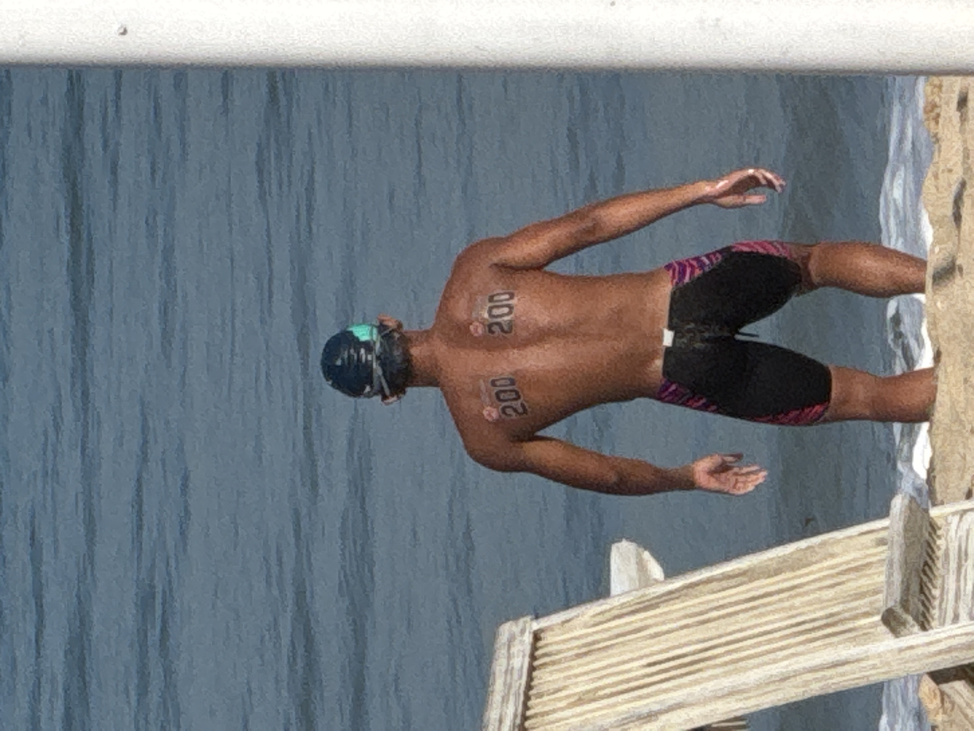 A person wearing a swim cap and goggles stands in the shallow water of Kuta Beach, Indonesia, facing away from the camera. The person's back is visible, with the number "200" tattooed in black on their left shoulder blade and a similar marking on their right shoulder blade. They are wearing dark swimming shorts with a pink and purple striped pattern on the sides. In the foreground, to the left, a portion of a light-colored wooden beach chair is visible. The background consists of the blue, rippling ocean. A white pole stands on the far right edge of the frame.
