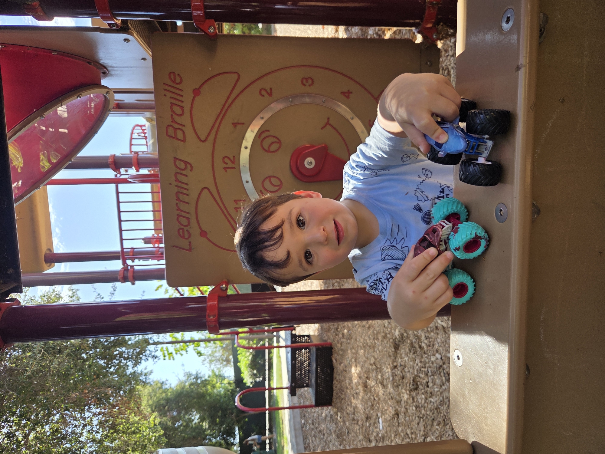 A young boy with dark hair and brown eyes sits on a playground structure, holding two toy monster trucks. He is wearing a light blue t-shirt with a graphic design. In his left hand, he holds a maroon and teal monster truck, while his right hand is positioned over a blue monster truck. The boy is looking towards the camera with a slight smile. Behind him, a large brown panel displays a clock face with numbers and a red hand. The playground is made of brown and red plastic, with metal poles supporting the structure. The ground is covered in wood chips. In the background, trees with green foliage and a clear blue sky are visible. A metal railing and a portion of another playground structure are also present. The location appears to be a public park or playground in Glendale, California, USA.