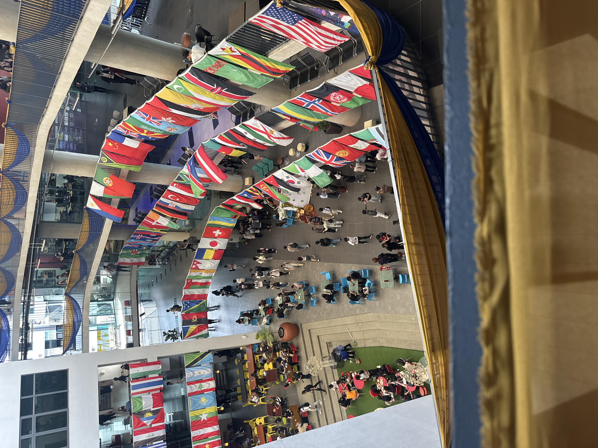 The image shows an indoor atrium decorated with rows of international flags. Tables and chairs are arranged in the open space, with people sitting and interacting. The view is from an elevated position, looking down upon the scene. The location is Kuala Lumpur, Malaysia.