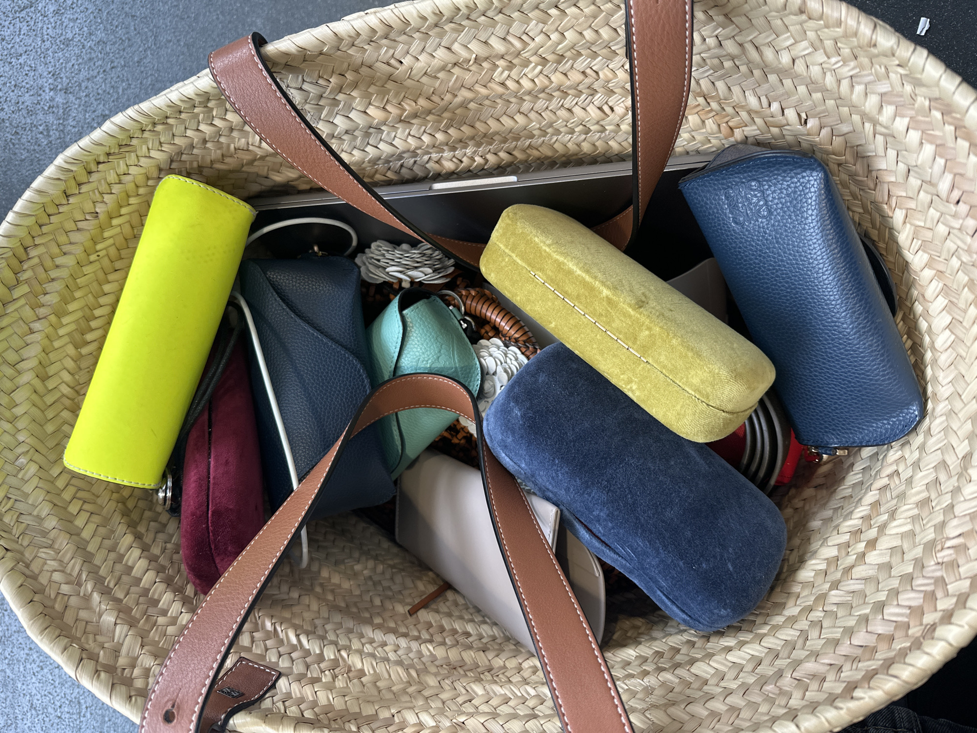 The image shows a straw tote bag filled with various colorful accessories and personal items. Inside, there are multiple eyeglasses cases in various colors (yellow, navy blue, light blue, dark velvet red, light brown), a light beige leather wallet, a black laptop, white paper flowers, and what appears to be a brown comb. The bag has brown leather straps. The background is a gray concrete surface. The location in the city of London, United Kingdom is not discernible from this image.