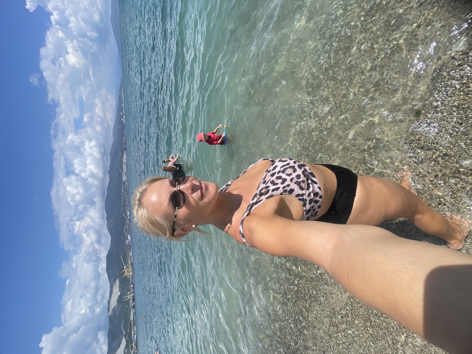 A woman is taking a selfie while standing in the shallow water of the sea. She has blonde hair, is wearing sunglasses and a leopard-print bikini top with black bottoms. In the background, there are other people in the water, a coastline with buildings, cranes, and mountains. The sea is clear and turquoise, and the sky is blue with white clouds. The location is likely the city of Novorossiysk, Russia, given the context.