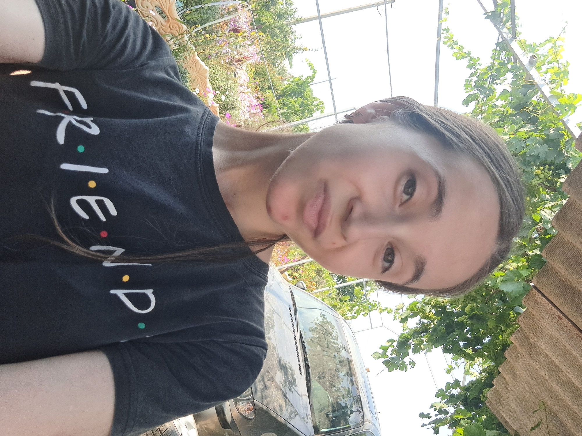 A young woman with long brown hair is taking a selfie outdoors. She is wearing a black t-shirt with the word "F.R.I.E.N.D.S." printed in white letters with colored dots. Her hair is partially pulled back, and she has a small earring in her left ear. Her face is turned slightly towards the camera, and she has a light expression.
The background shows a car, some green foliage from a vine or tree overhead, and a colorful garden with pink and white flowers. The setting appears to be a rural or suburban area. Based on the context provided, the location is Vypasne, Ukraine. The image is taken during daylight, with natural light illuminating the scene.