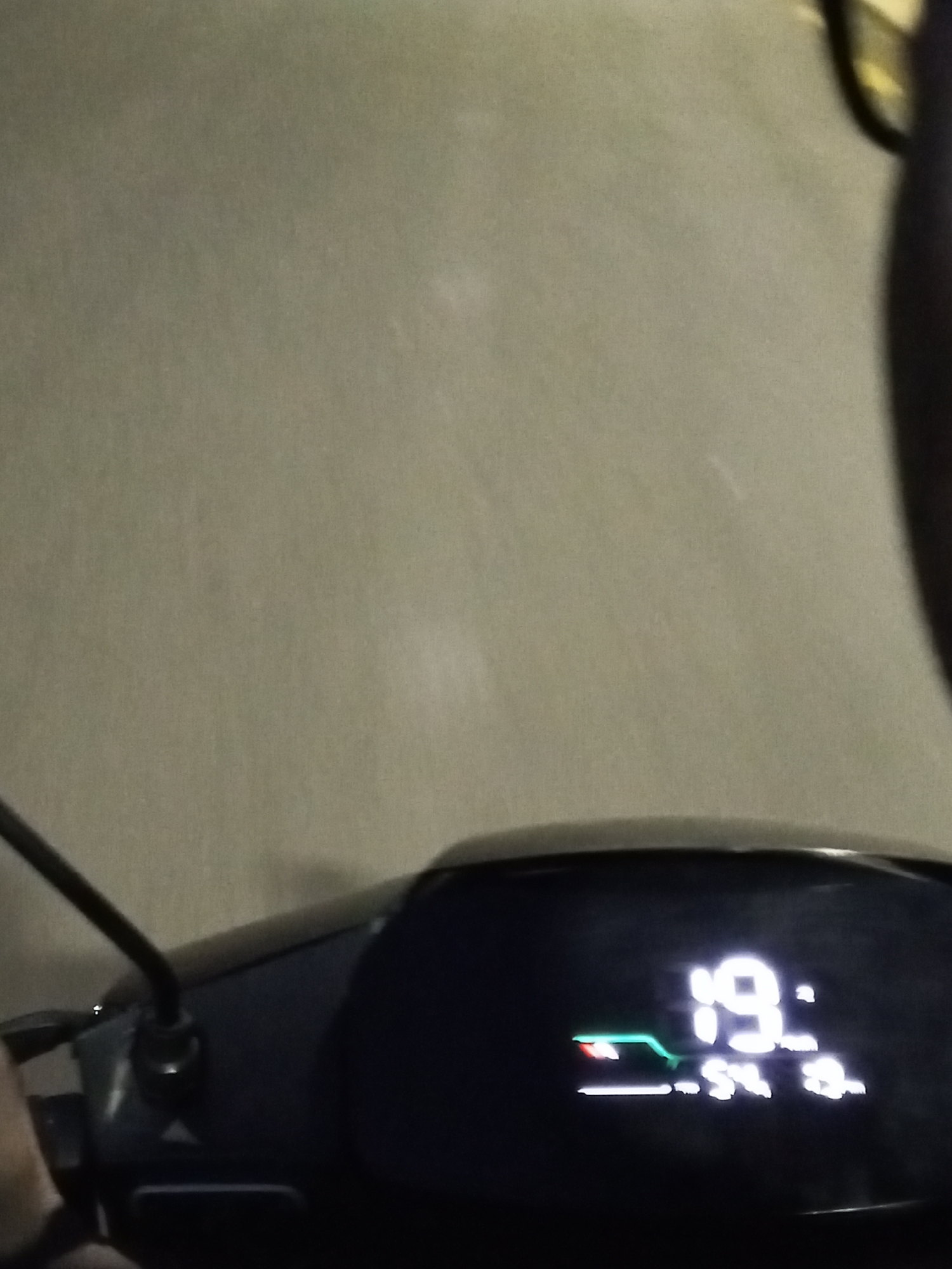 The image shows a close-up of a digital display on a vehicle, likely a scooter or motorcycle. The display shows a speed of "19:" which likely represents 19 km/h. Below that, the display shows "-54 3". The display is illuminated and set against a dark background. In the background, there is a textured, light-colored surface, possibly a wall or road. The image appears to be taken at night or in low light conditions. There is no identifiable information that places this image in Jos, Nigeria.