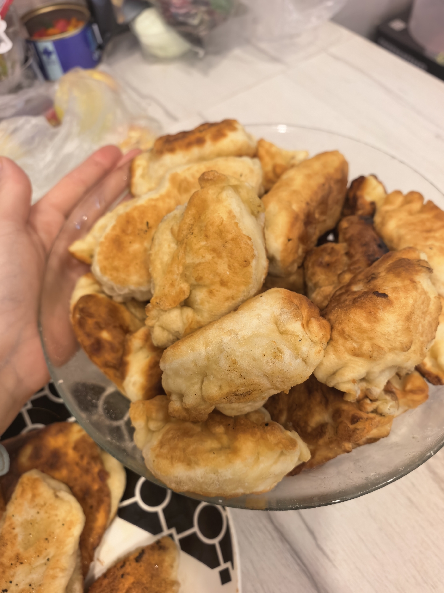 A close-up, overhead shot shows a clear glass bowl overflowing with golden-brown fried pastries, possibly chebureki or similar fried dumplings. A hand with a light skin tone is visible on the left side of the frame, holding the bowl. The pastries have a slightly uneven, crimped edge and appear to be filled. Some are stacked on top of others, creating a dense arrangement within the bowl. The background is blurred but suggests an indoor setting with a light-colored surface, possibly a table, and some indistinct items on a shelf or counter. A black and white patterned surface is visible beneath the bowl at the bottom of the image. The lighting is warm, highlighting the fried texture and color of the food. The image composition focuses entirely on the pastries and the hand holding the bowl, with no discernible elements that would indicate a specific location in Sterlitamak, Russia.