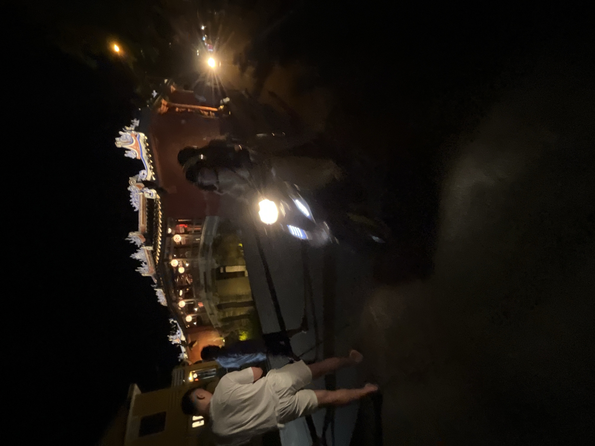The image depicts a night scene in Hội An Ward, Vietnam, likely near a river or canal area with traditional architecture. In the foreground, a person in a white t-shirt and white shorts stands on a paved surface. To their left and slightly behind, another person in a blue shirt and dark shorts is visible. A motorcycle with its headlights on is in the mid-ground, with two individuals seated on it. The rider is wearing a light-colored outfit, and the passenger is wearing a darker, possibly hooded garment. Behind them, a building with illuminated lanterns and ornate roof details is visible, characteristic of Hoi An's ancient town. The lighting suggests it is nighttime, with artificial lights from the buildings, lanterns, and vehicle headlights illuminating the scene. The atmosphere is dark, with the illuminated areas standing out against the blackness of the night sky and unlit portions of the environment.