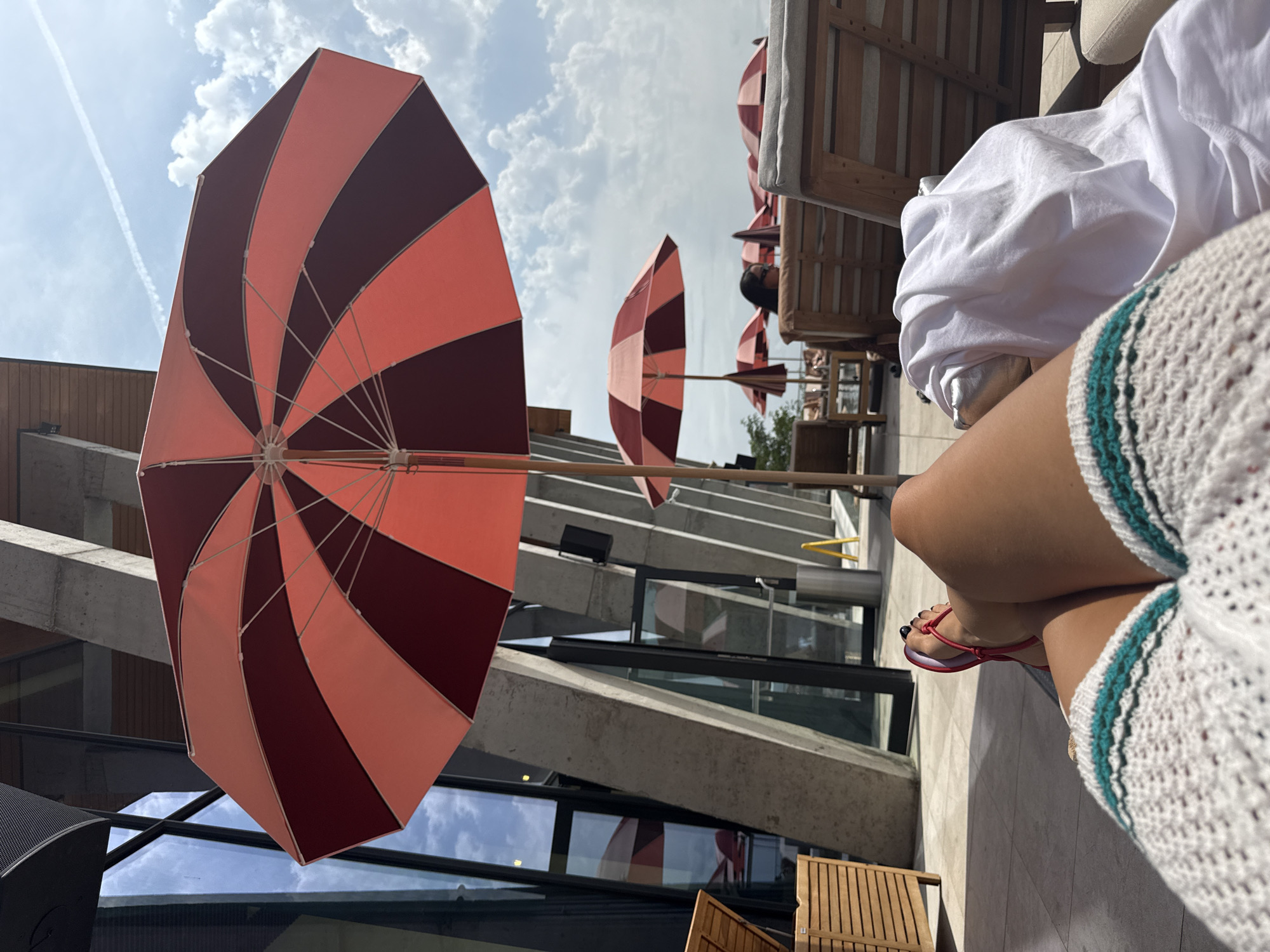 A person is relaxing in a lounge chair by a pool on a sunny day. The person is wearing a white crocheted cover-up and red flip-flops. A large red and white striped umbrella is open overhead, providing shade. In the background, there are other lounge chairs and umbrellas, as well as a modern building with large glass windows. The scene appears to be a resort or a hotel with a pool area. There is no indication that this location is in Sighnaghi, Georgia.