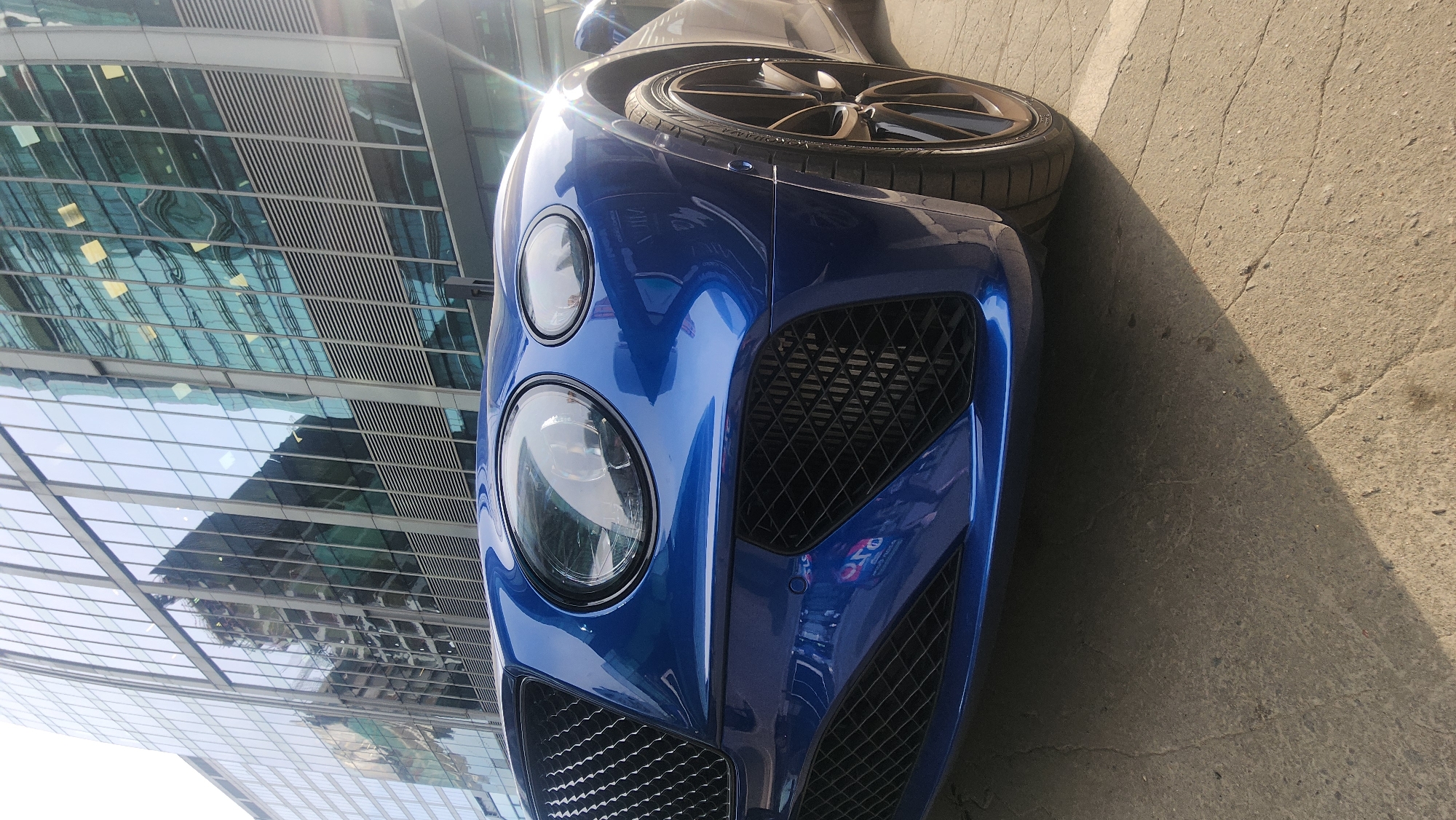 A blue Bentley automobile is parked on a concrete surface adjacent to a modern glass-walled building, reflecting the city skyline. The car's front end, including the headlights, grille, and front tire, are prominently visible. The photograph appears to have been taken in the Moscow International Business Center, also known as Moscow City, Russia, due to the distinctive architectural style of the buildings in the background. The overall lighting suggests it is daytime.