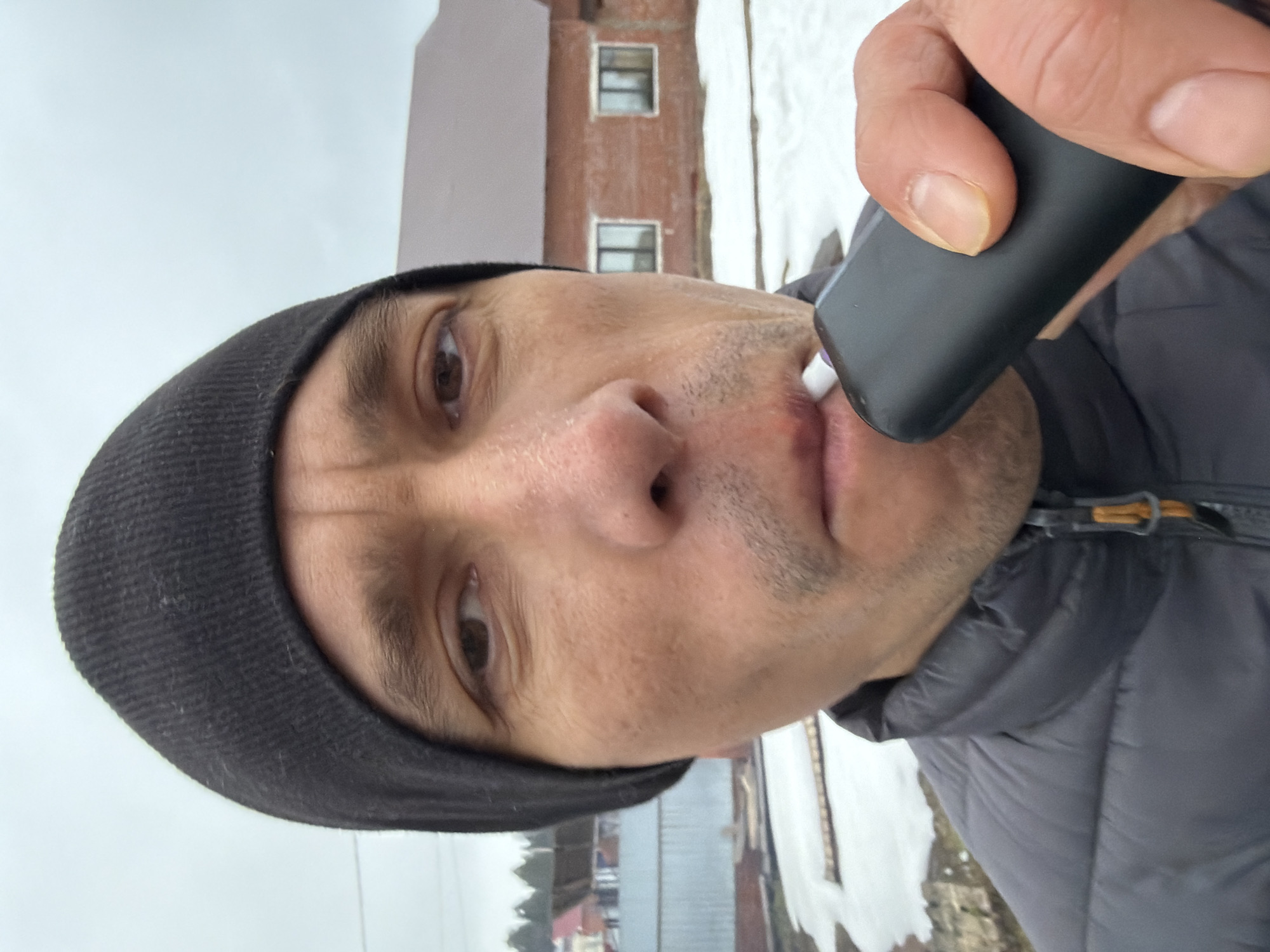 A man is holding a black electronic device to his mouth, possibly vaping, while standing outside in what appears to be a snowy or cold environment. He is wearing a black beanie and a dark puffer jacket.
In the background, there is a building with red brick exterior and windows. The presence of snow on the ground and on the building suggests it is winter or a cold season. Other buildings and trees are visible further in the background, indicating a residential area or town.
While it's impossible to determine the exact location in Chaikovsky, Russia, without more information, the architecture and environment suggest a residential area within the city.