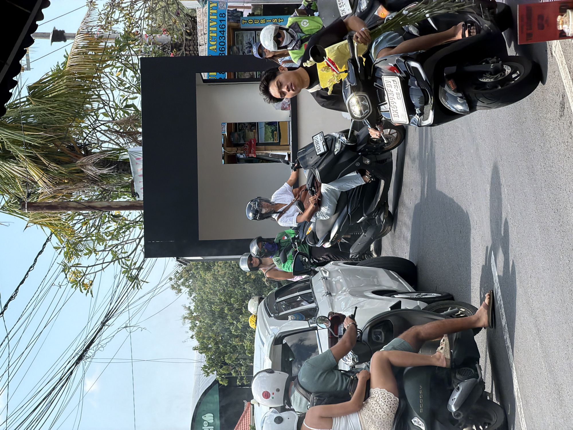 The image depicts a vibrant street scene in North Kuta, Indonesia, under bright, clear daylight, suggesting midday or early afternoon with strong sun casting sharp shadows. The setting is a busy road, likely experiencing traffic, filled with numerous people primarily riding scooters and motorbikes, alongside a white SUV car.
In the immediate foreground, a person with bare feet is seated on a black "SCOOPY" brand scooter. Adjacent, another person on a black "HONDA" scooter with license plate "DK 5993 FDV" (valid until "08-25") is holding a bundle of green plants. Further back, a man in a black t-shirt, holding a yellow bag and also on a scooter, looks directly at the camera. Another scooter with license plate "DK 5196 FFV" (valid until "07-24") is visible in the line of traffic. Many individuals are wearing helmets, and some have face masks. A person in a green vest and white cap, possibly a ride-hailing driver, is also seen.
The background features commercial buildings, including one with a dark facade. Overhead, a dense entanglement of power lines stretches across a clear blue sky, interspersed with lush green palm trees and other foliage.
Visible text includes: "DK 5993 FDV" and "08-25" on a scooter license plate, "DK 5196 FFV" and "07-24" on another, "HONDA" on a scooter, "CANGGU" on a scooter's fender, and "SCOOPY" on a scooter's side. A blue sign reads "ACTIVITY TOURIST" (with "TOURIST" misspelled) and "6034618". Parts of a red sign with "THE CANGGU" and a green sign with an elephant graphic, possibly saying "ELEPHANT", are also visible.