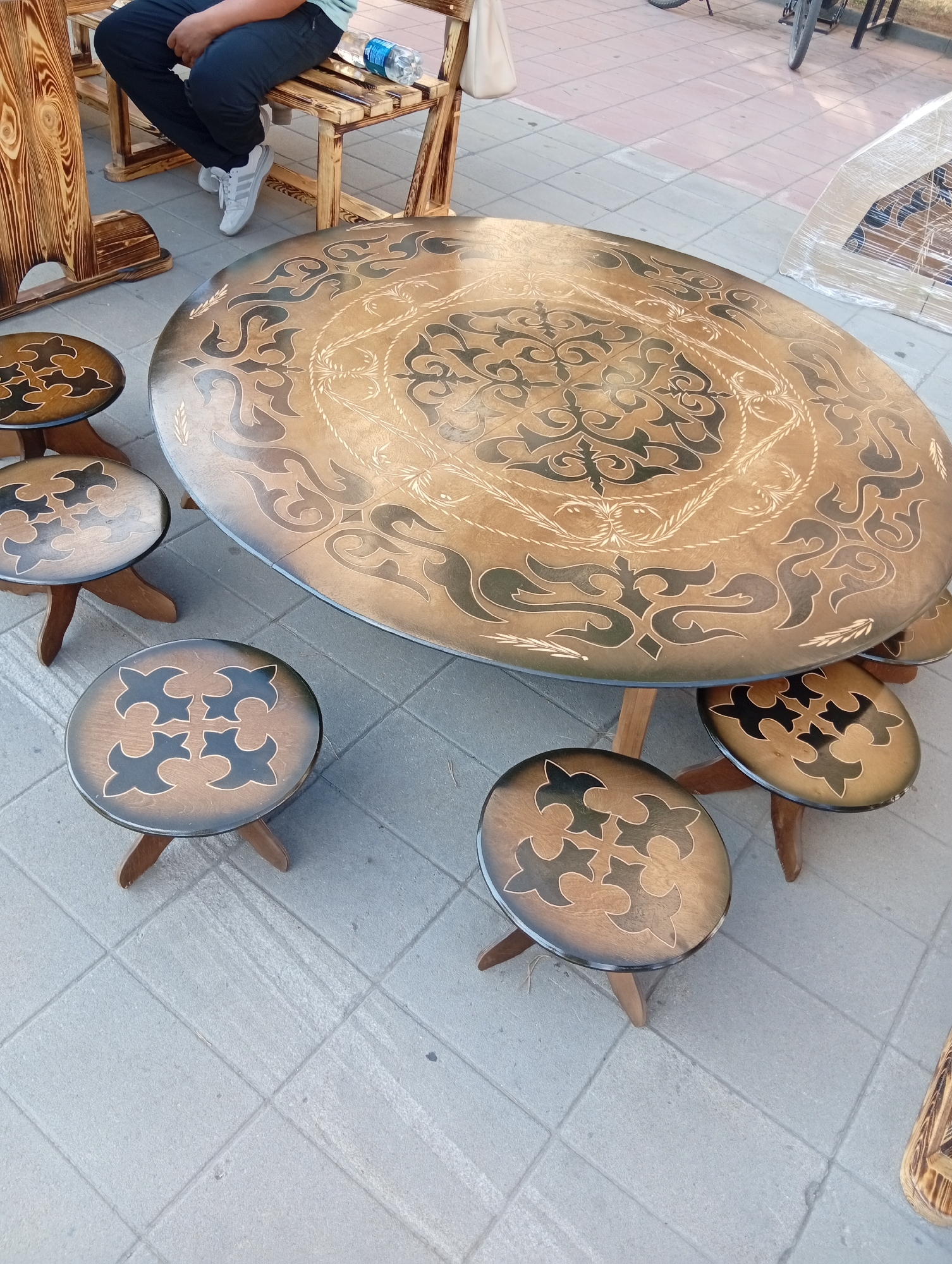 The image depicts an outdoor market scene, likely in Kostanay, Kazakhstan, showcasing a set of handcrafted wooden furniture. In the foreground, a large, oval-shaped wooden table is prominently displayed. Its surface is decorated with intricate, dark brown and light brown geometric patterns, possibly inspired by traditional Kazakh designs. Surrounding the table are several small, round wooden stools, each with a similar decorative pattern on its top. The furniture appears to be new and is arranged on a paved outdoor area, with square grey tiles forming the ground.
In the background, a person is seated on a wooden bench, wearing dark trousers and white sneakers. The person's hands are resting on their lap, and a blue plastic bottle of water is visible on the bench next to them. To the left of the bench, a portion of another wooden item, possibly a display stand or part of a larger furniture piece, with a distinct wood grain pattern is visible. Further in the background, parts of a bicycle and a wrapped item covered in plastic sheeting suggest the presence of other market stalls or goods. The overall impression is that of a vibrant outdoor marketplace.