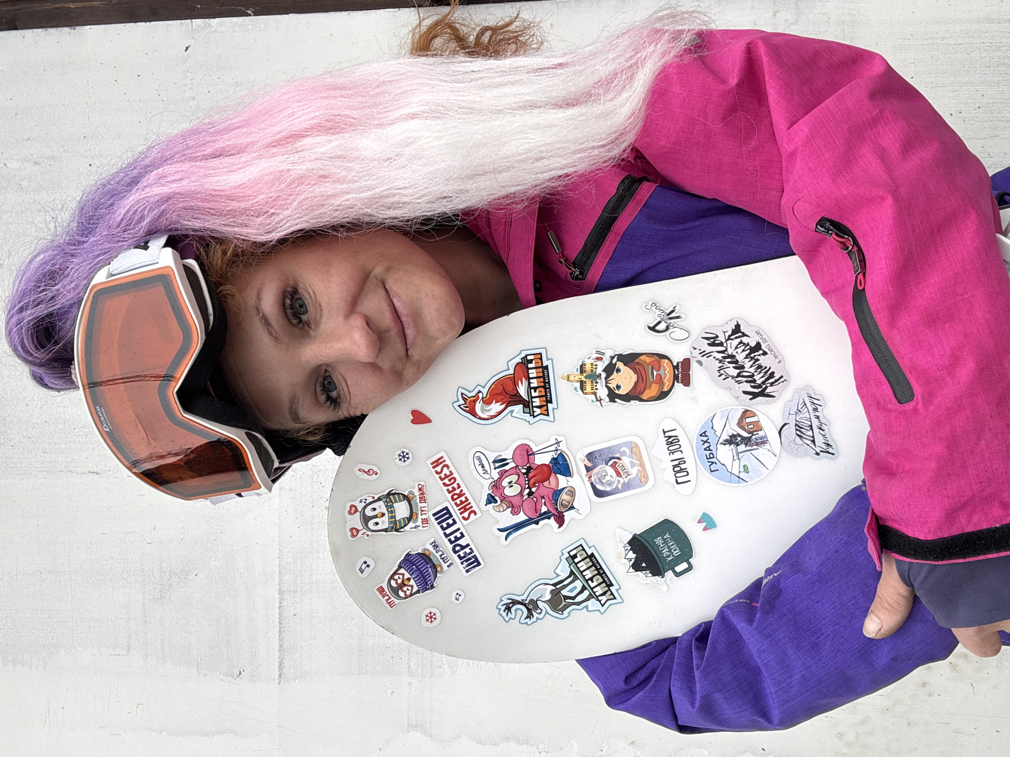 A smiling person is depicted posing with a white snowboard, indicating an enthusiasm for winter sports. They have distinctive long hair with an ombre style, transitioning from purple at the roots to pink and then white. The individual wears a vibrant fuchsia-pink winter jacket with purple details and has orange-tinted ski goggles, marked "DOUBLEFLEX", resting around their neck/head.
The white snowboard is adorned with numerous colorful stickers, many of which reference popular Russian ski resorts and mountain regions. Visible text on these stickers includes "ШЕРЕГЕШ SHEREGESH", "ХИБИНЫ POWER OF NATURE", "КРАСНАЯ ПОЛЯНА", "ГОРЫ ЗОВУТ" (The mountains call), "ГУБАХА", and "Домбай". Other stickers show cartoon characters, a fox, penguins, and phrases such as "ПУХЛЯШ", "В ПУХЛЯКЕ ГДЕ ТУТ ДВИЖ?", "Куискумчорр", and "Кабины в моей душе".
The person looks directly at the camera with a happy and relaxed expression. The bright, even lighting suggests it is daytime. The background is a plain, light-colored surface, possibly snow or a wall, emphasizing the subject and their personalized snowboard. The overall impression is one of joy and passion for snowboarding and exploring various winter destinations, consistent with the location context of Chaikovsky, Russia.