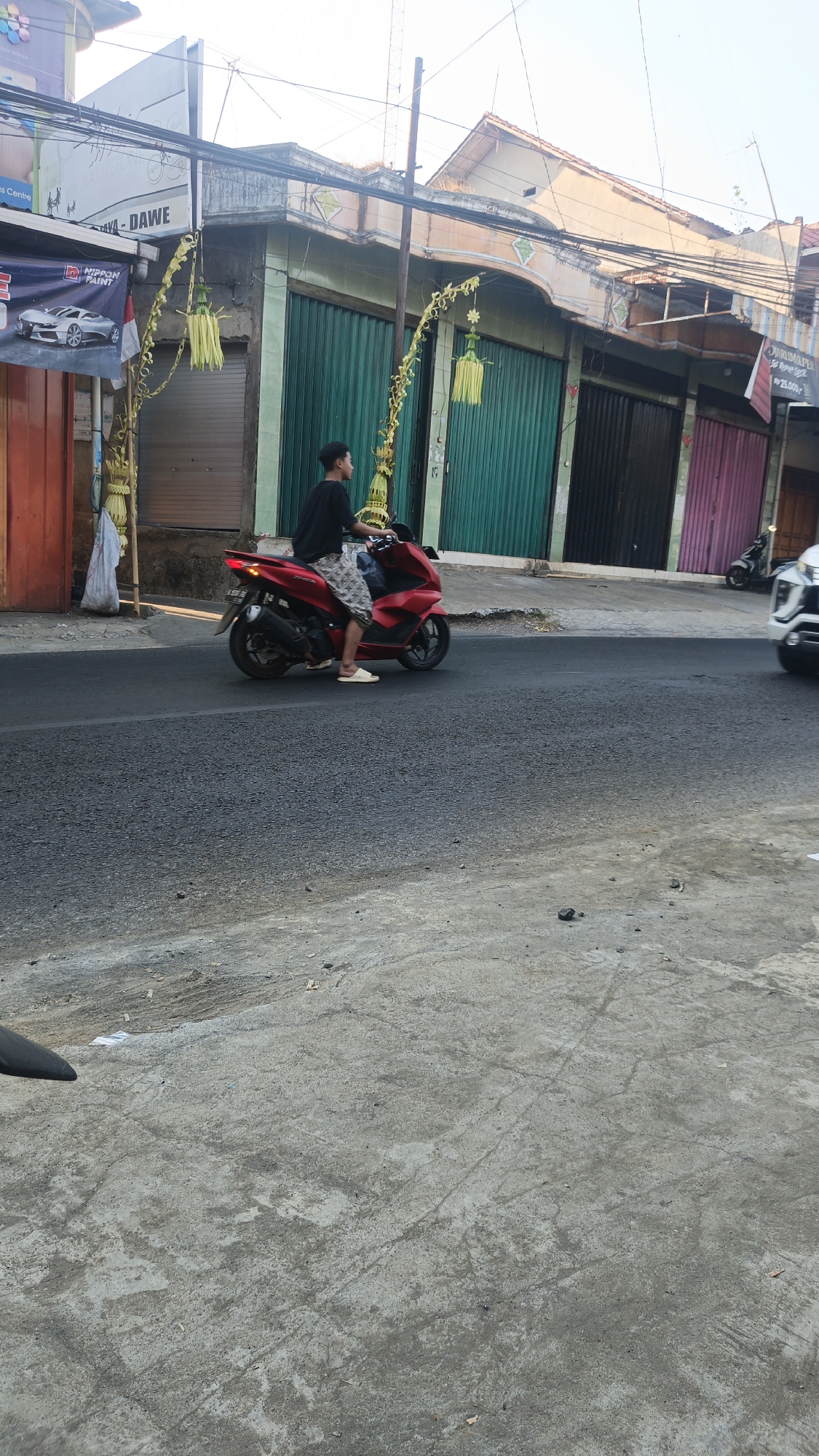 An overhead, slightly angled, eye-level view captures a person riding a red motorcycle on a paved road in Dawe, Indonesia. The rider, wearing a black t-shirt and patterned shorts, is seated on the motorcycle, which is positioned in the middle of the road. The motorcycle's rear brake light is illuminated, suggesting it is in motion. The background features a row of buildings with green and pink roll-up doors. Festive yellow decorations, possibly for a local celebration, are visible along the street. The road surface is asphalt, with a lighter-colored concrete area in the foreground. The sky is clear and bright, indicating daylight.