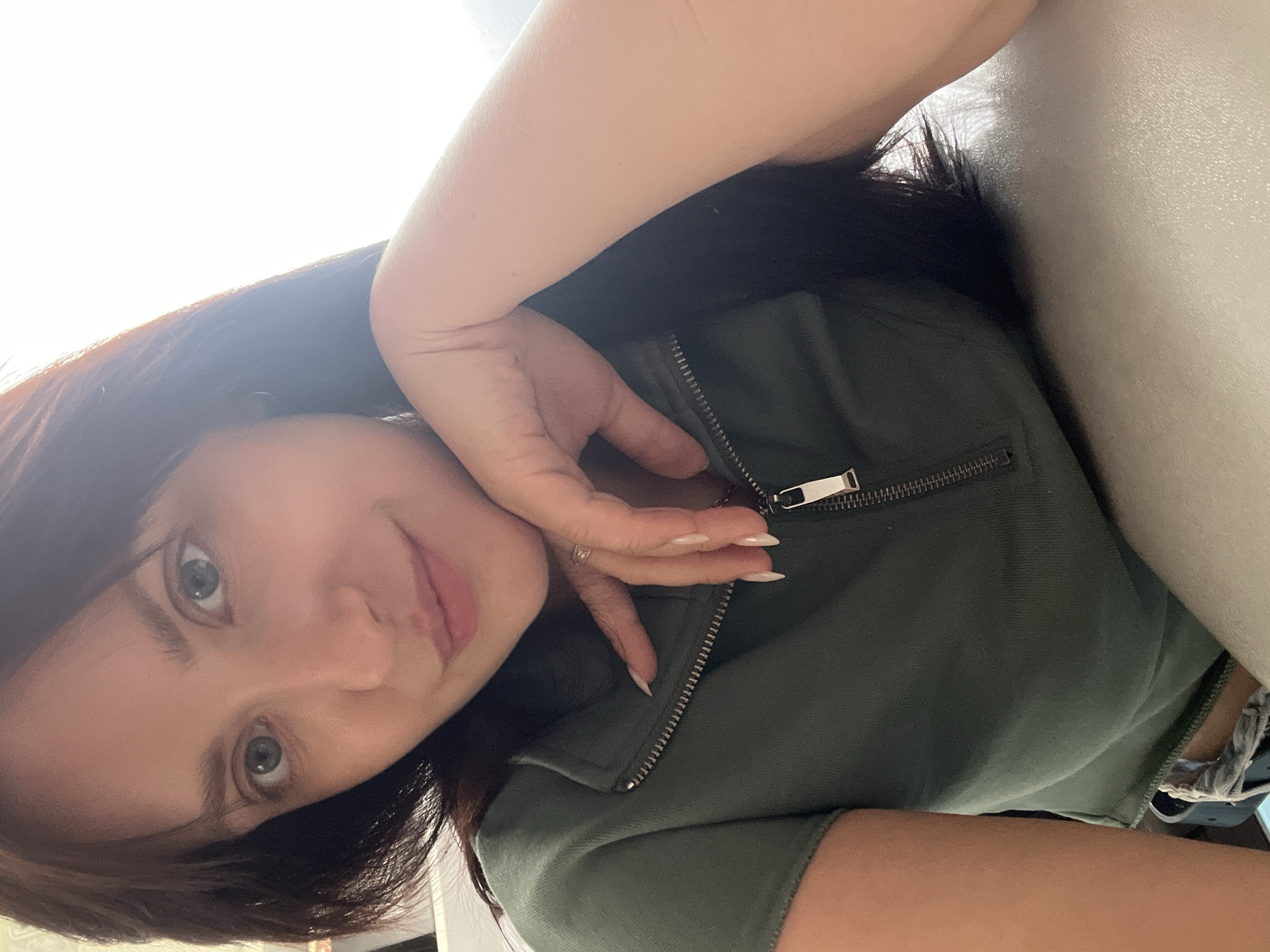 A woman with dark hair and fair skin is looking at the camera with a slight smile. She is wearing a dark green, zippered top. Her left hand is near her face. The background appears to be an interior space. There is no indication of the location being Марковский, Russia.